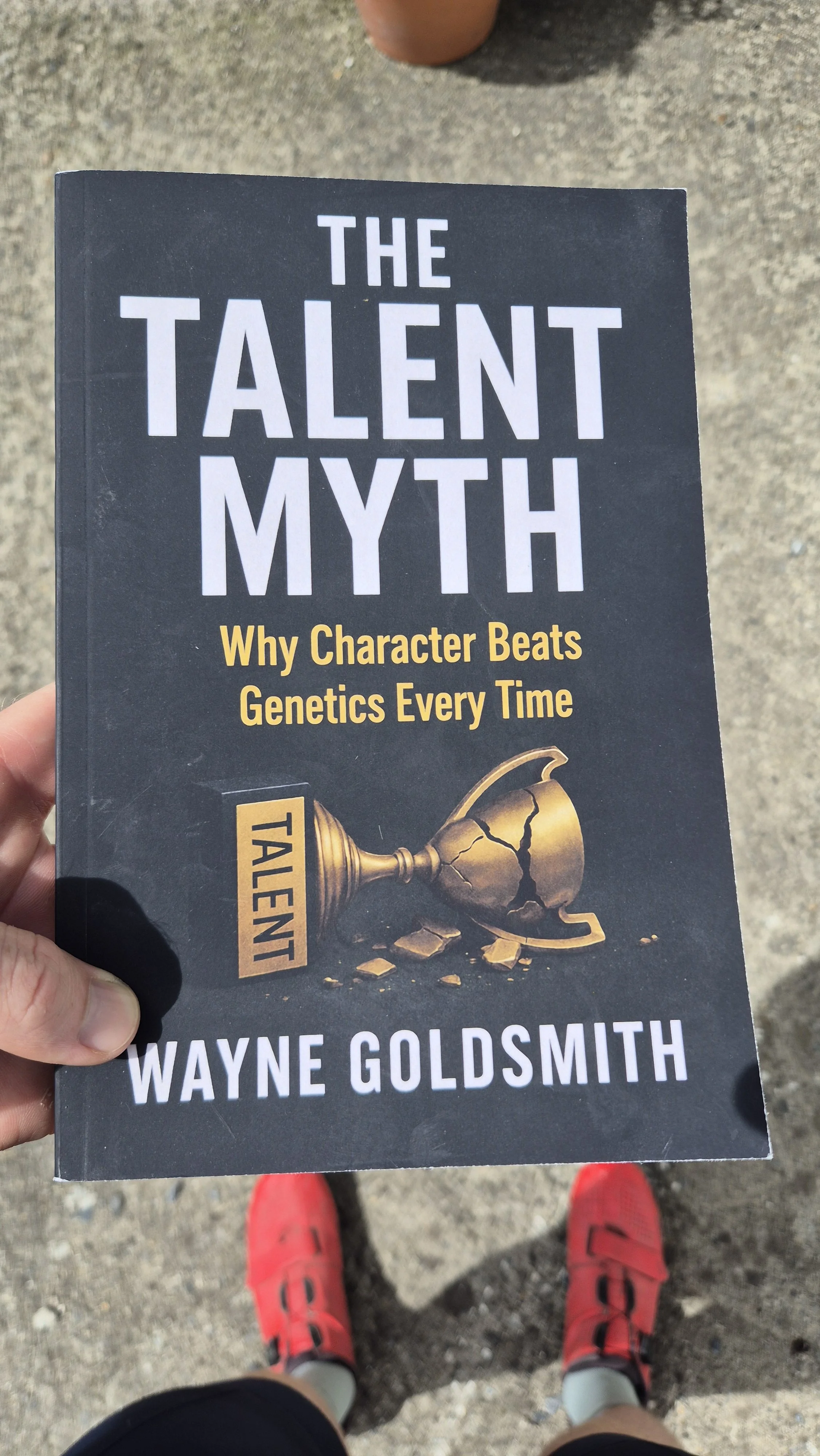 A person holding a book titled "The Talent Myth" by Wayne Goldsmith, with a cracked trophy on the cover and the subtitle "Why Character Beats Genetics Every Time," outdoors on a gravel surface.