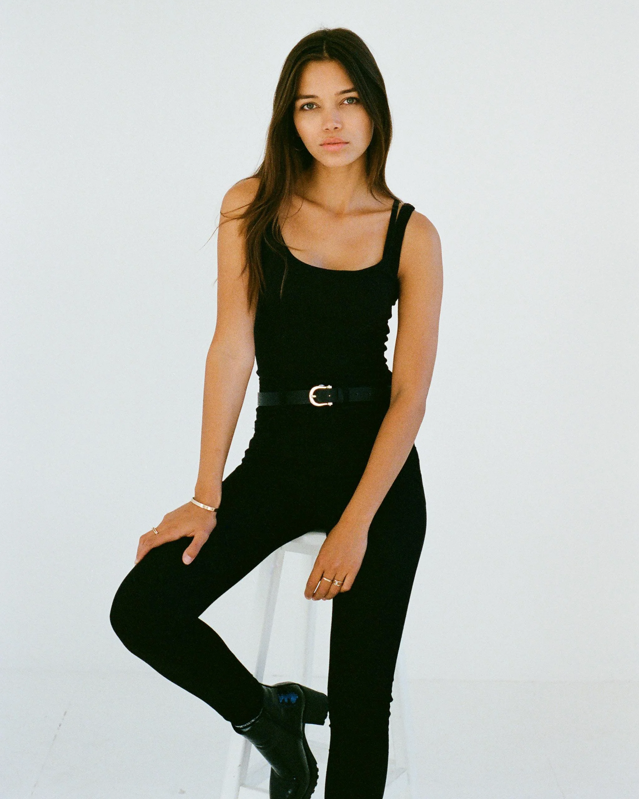 A woman sitting on a white chair with one leg crossed over the other, wearing a black sleeveless top and black pants, with long dark hair, in front of a plain white background.