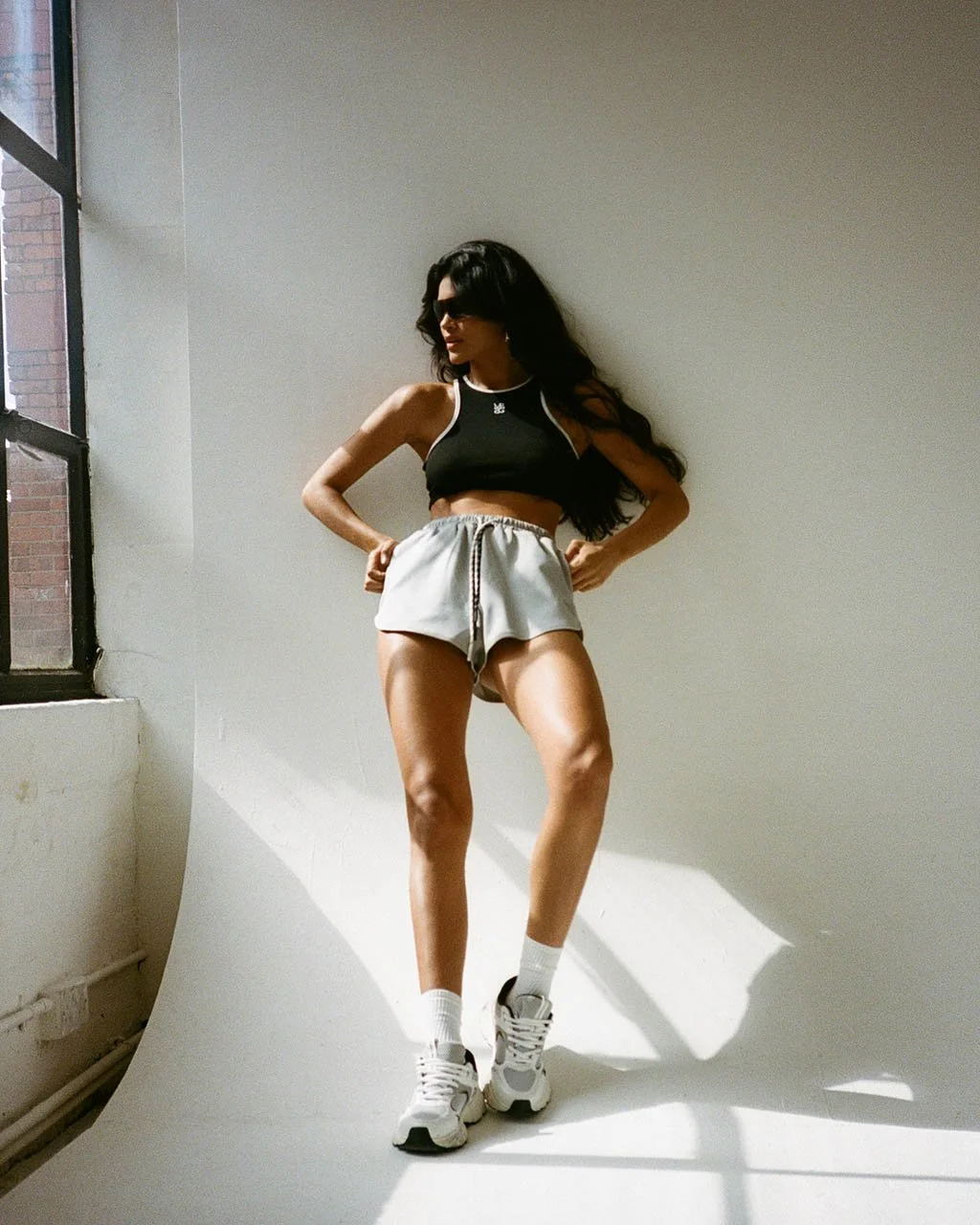 A woman with long dark curly hair wearing sunglasses, black crop top, grey shorts, white socks, and athletic shoes, standing inside near a window with sunlight streaming in.