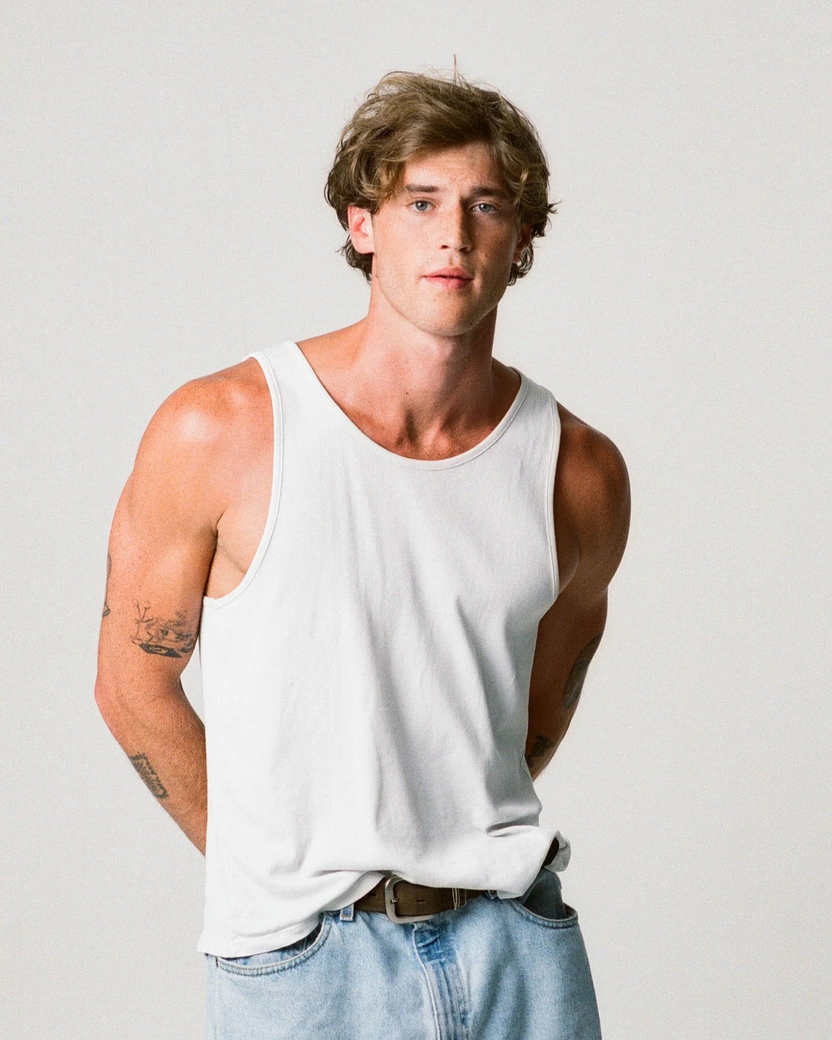 A young man with wavy brown hair, wearing a white tank top and light blue jeans, stands against a plain white background.