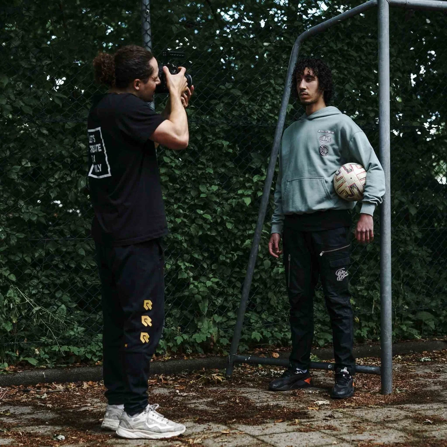 Twee mannen, waarvan één een camera vasthoudt en de ander tegen een voetbaldoel staat met een voetbal onder zijn arm. Omringd door groen gebladerte.