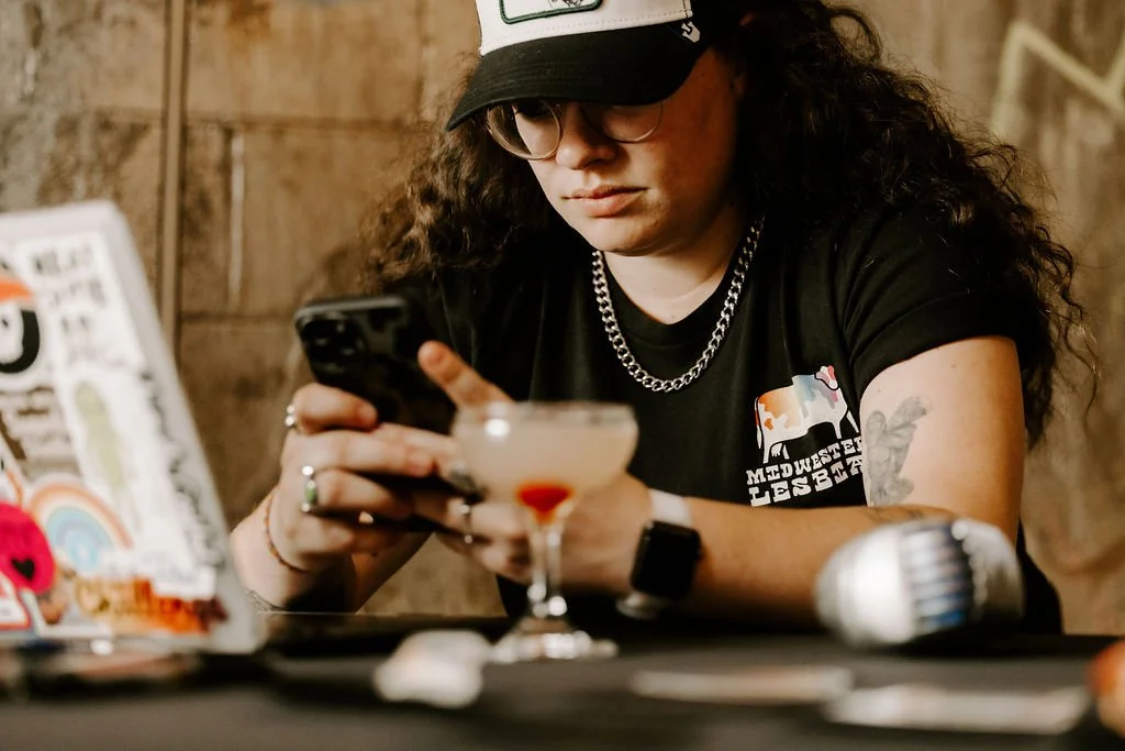 Person wearing a cap and glasses using a smartphone, seated at a table with a cocktail, a laptop, and a microphone. They are wearing a shirt with an illustration and text on it.
