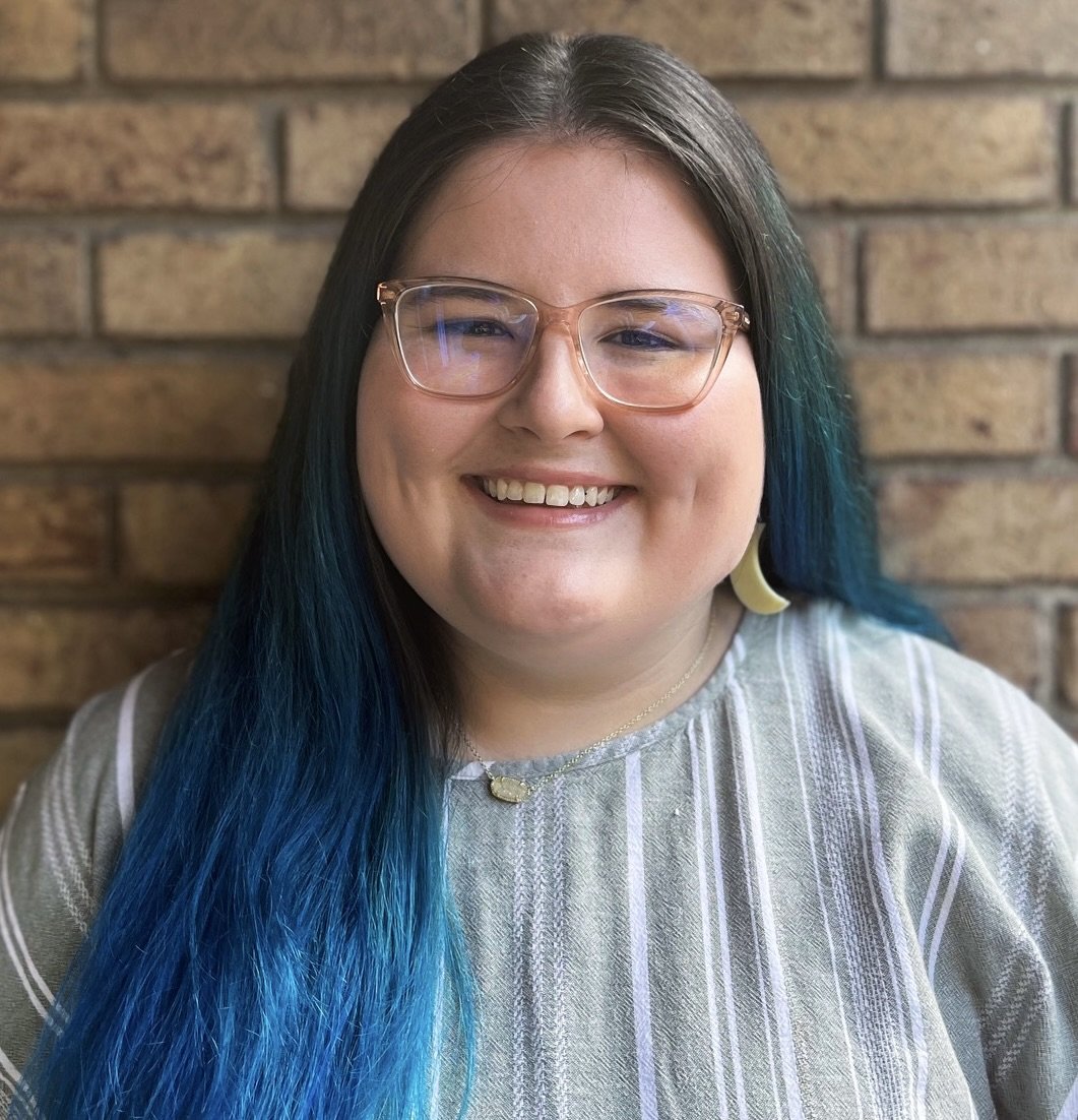 Smiling person with glasses and long blue hair, wearing a striped shirt, in front of a brick wall.