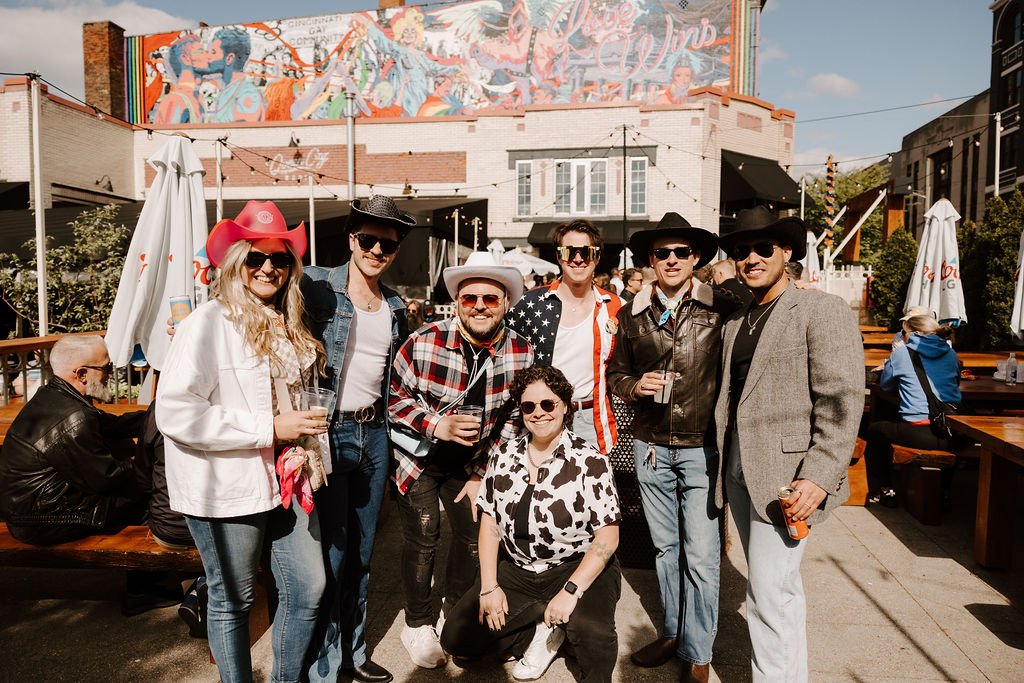 Group of people outdoors wearing cowboy hats and casual attire in front of a colorful mural.