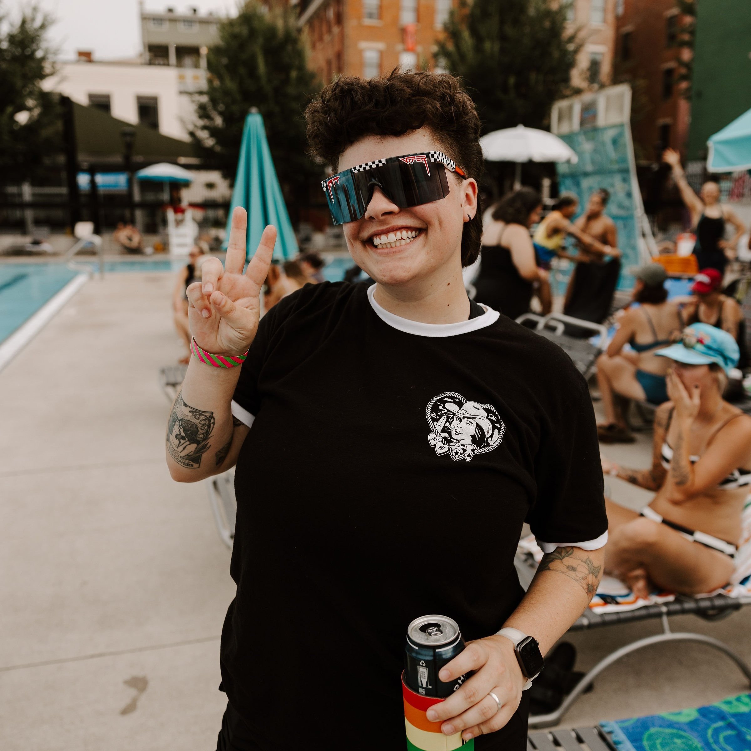 Person at a pool party wearing sunglasses and a black T-shirt, holding a drink in a rainbow koozie, making a peace sign with their hand. Other people relax and socialize by the poolside.