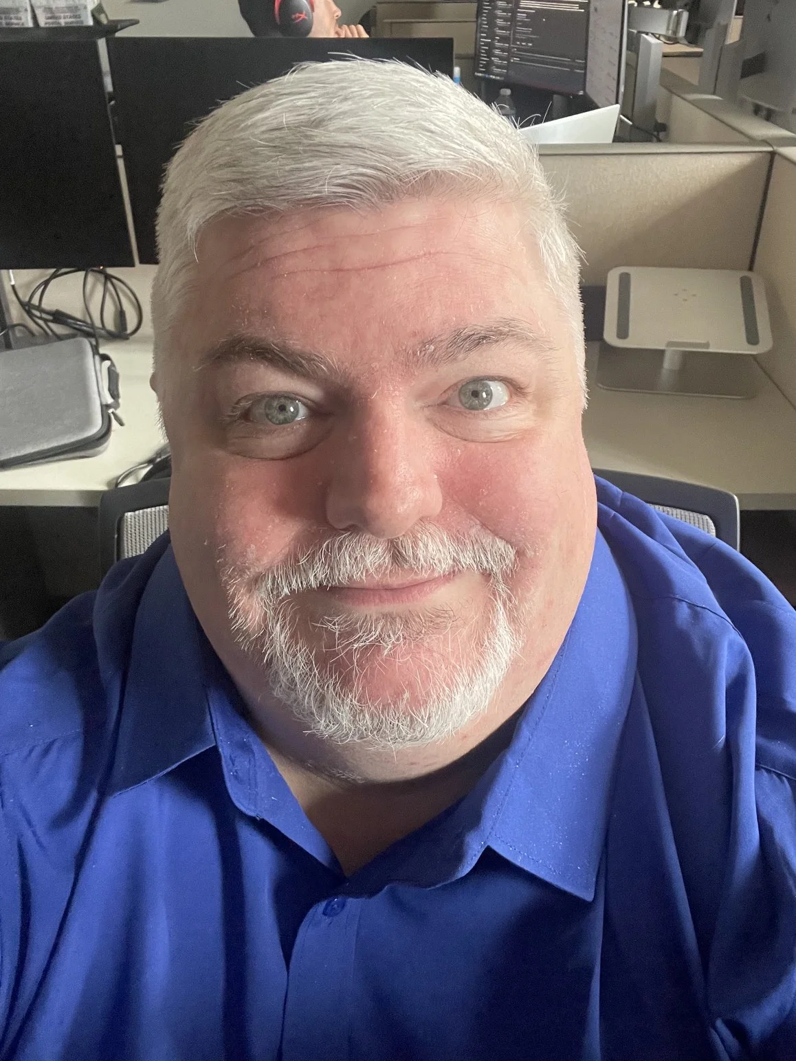 A cheerful middle-aged man with gray hair and beard taking a selfie in an office cubicle. He is wearing a blue button-up shirt, and in the background, there are computer monitors and a person with headphones working at a desk.