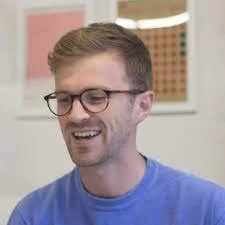 A man with glasses and short brown hair, smiling and wearing a blue shirt, sitting indoors.