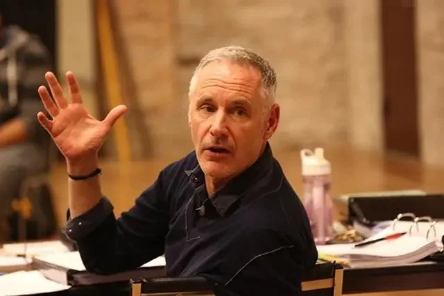 A young man with short gray hair, wearing a black shirt, sitting at a table with papers and a pink water bottle, looking at the camera and gesturing with his right hand.