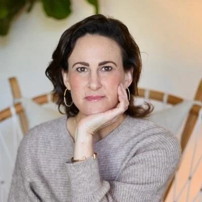 Person sitting in a chair with a neutral expression, hand resting on chin, wearing a gray sweater and hoop earrings.