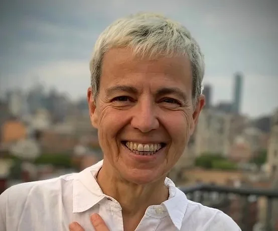 Smiling middle-aged woman with short gray hair standing outdoors with a cityscape in the background.