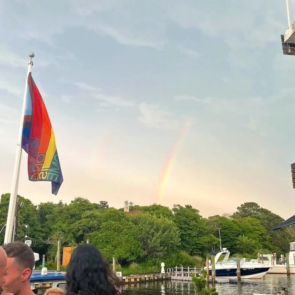 A harbor scene with boats docked, a rainbow in the sky, and a rainbow flag in the foreground.
