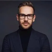 Portrait of a man with glasses, wearing a dark suit and black turtleneck, against a dark background.