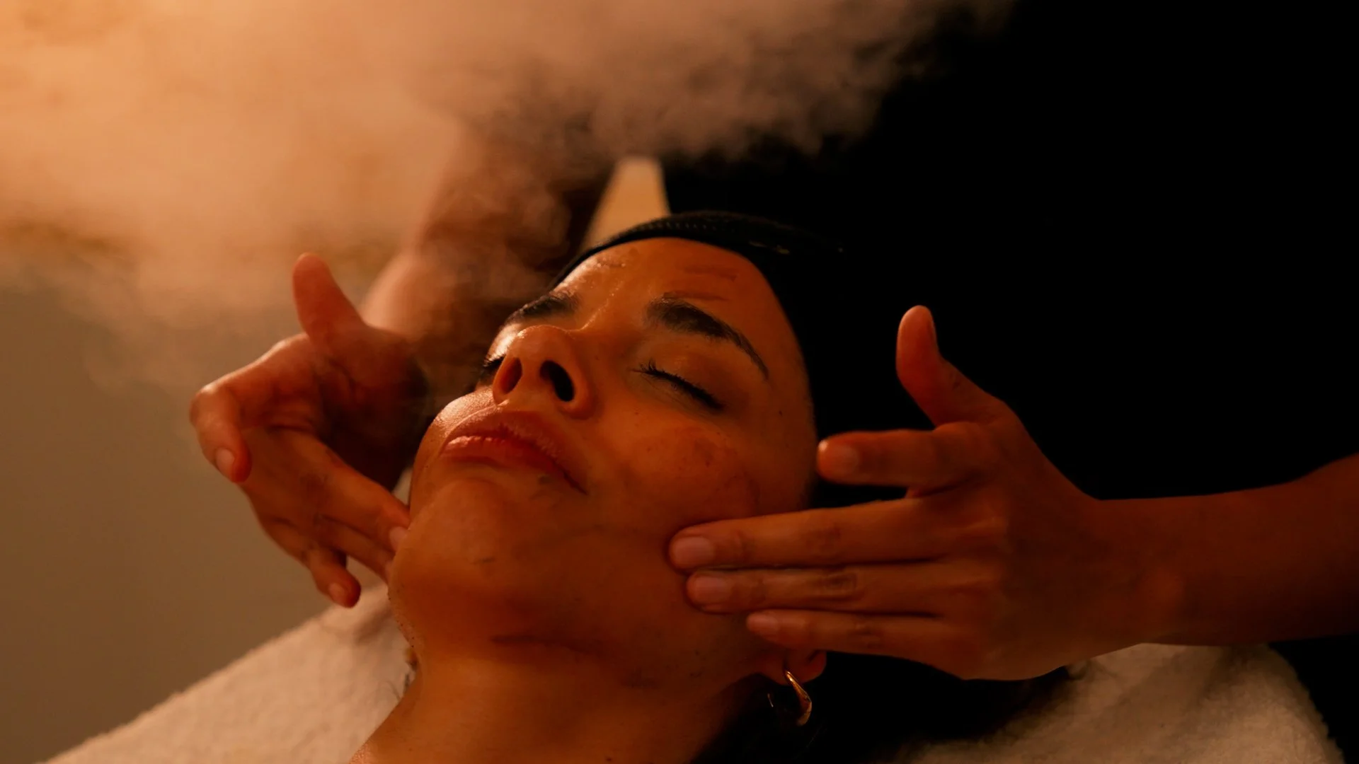A person lying face up with their eyes closed, receiving a facial treatment from a practitioner whose hands are gently massaging the person's face in a dimly lit room.
