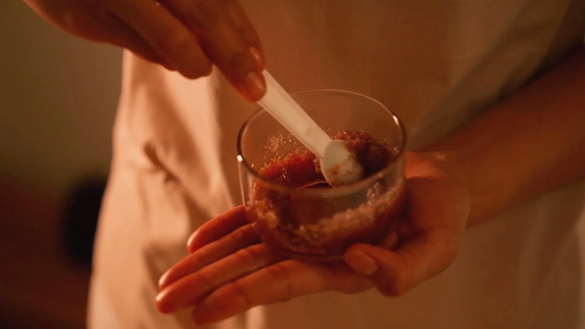 A person is stirring a glass of a dark red, likely alcoholic, beverage with a white straw. The image is shot in warm lighting, and the person's hands and part of their beige or light-colored clothing are visible.