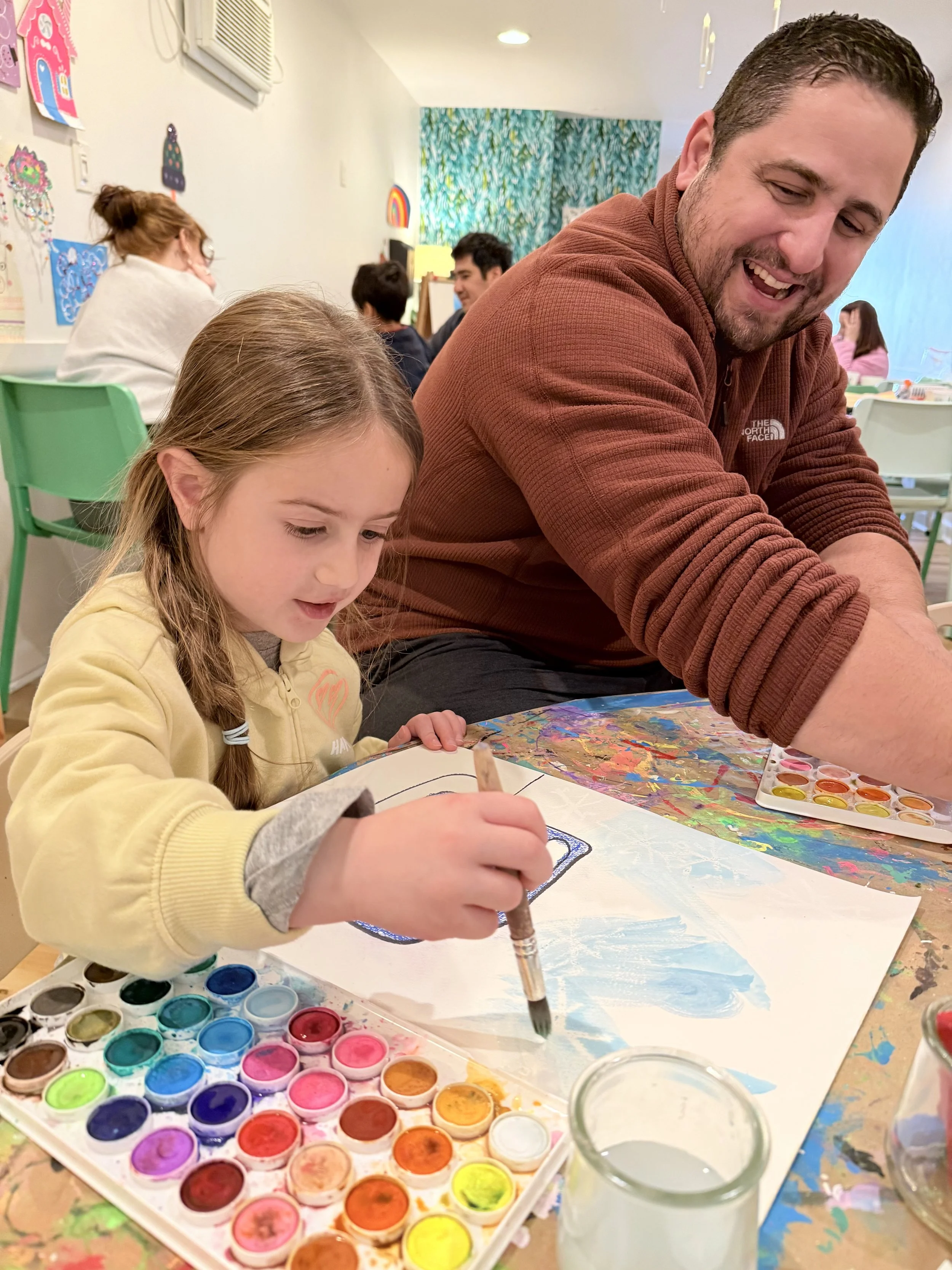 A young girl and man painting at a colorful art table in a classroom. The girl is focused on her watercolor painting, while the man watches and smiles.