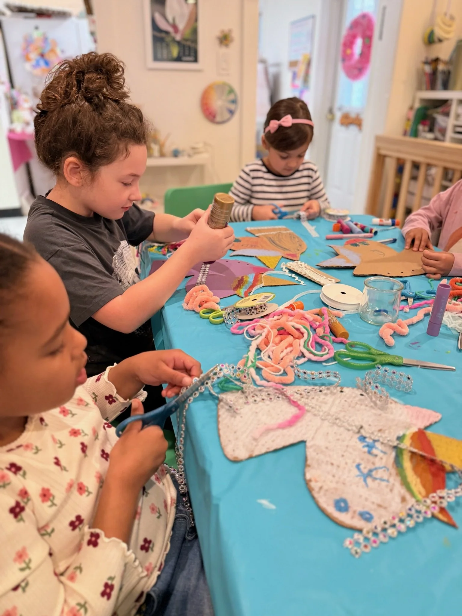 Two young girls sitting at a table with colorful art supplies and decorated foam shapes, engaging in craft projects, with colorful children's artwork displayed on the wall behind them.