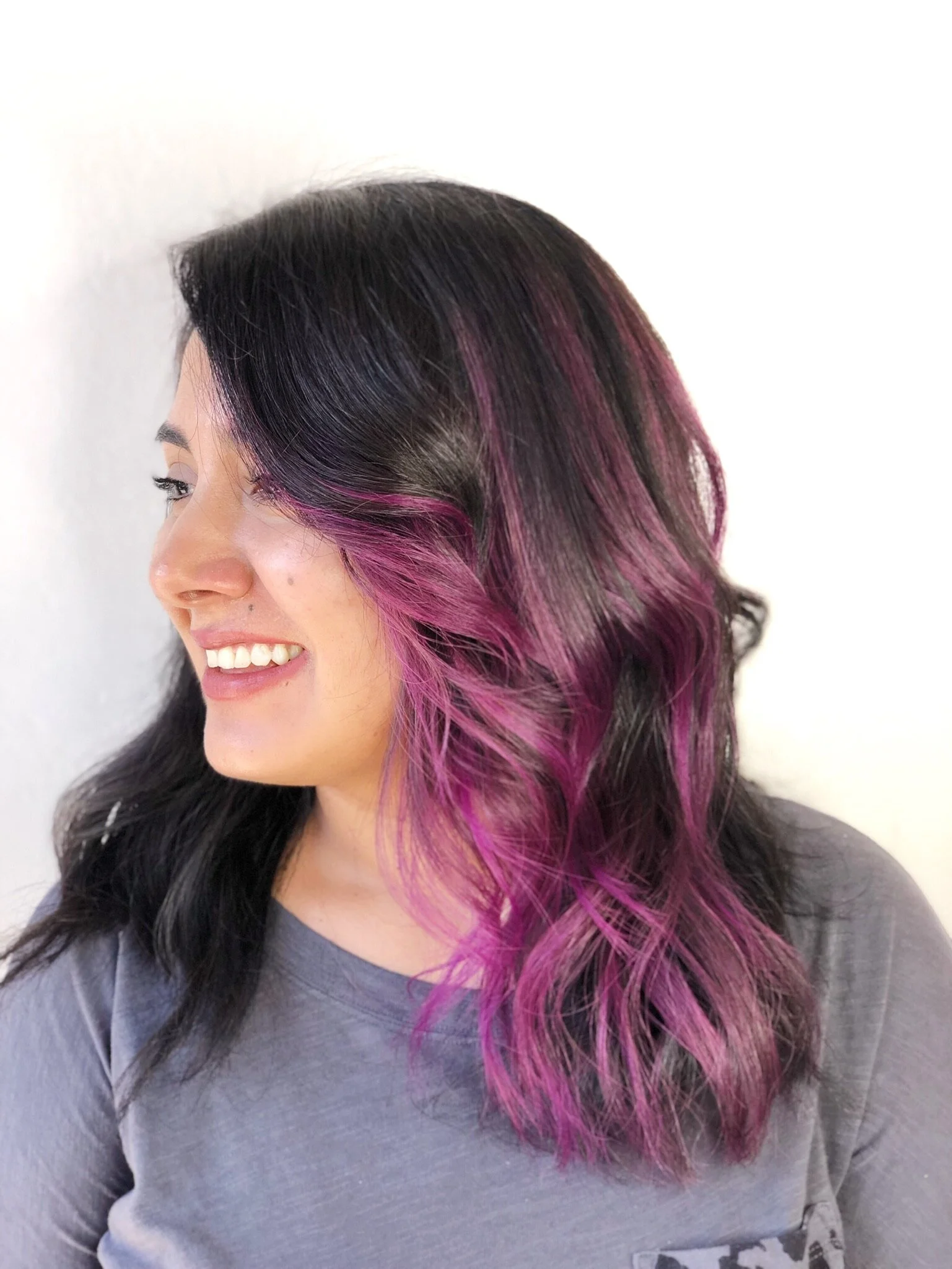 Woman with long, wavy black hair with pink highlights on the ends, smiling and looking to the side.