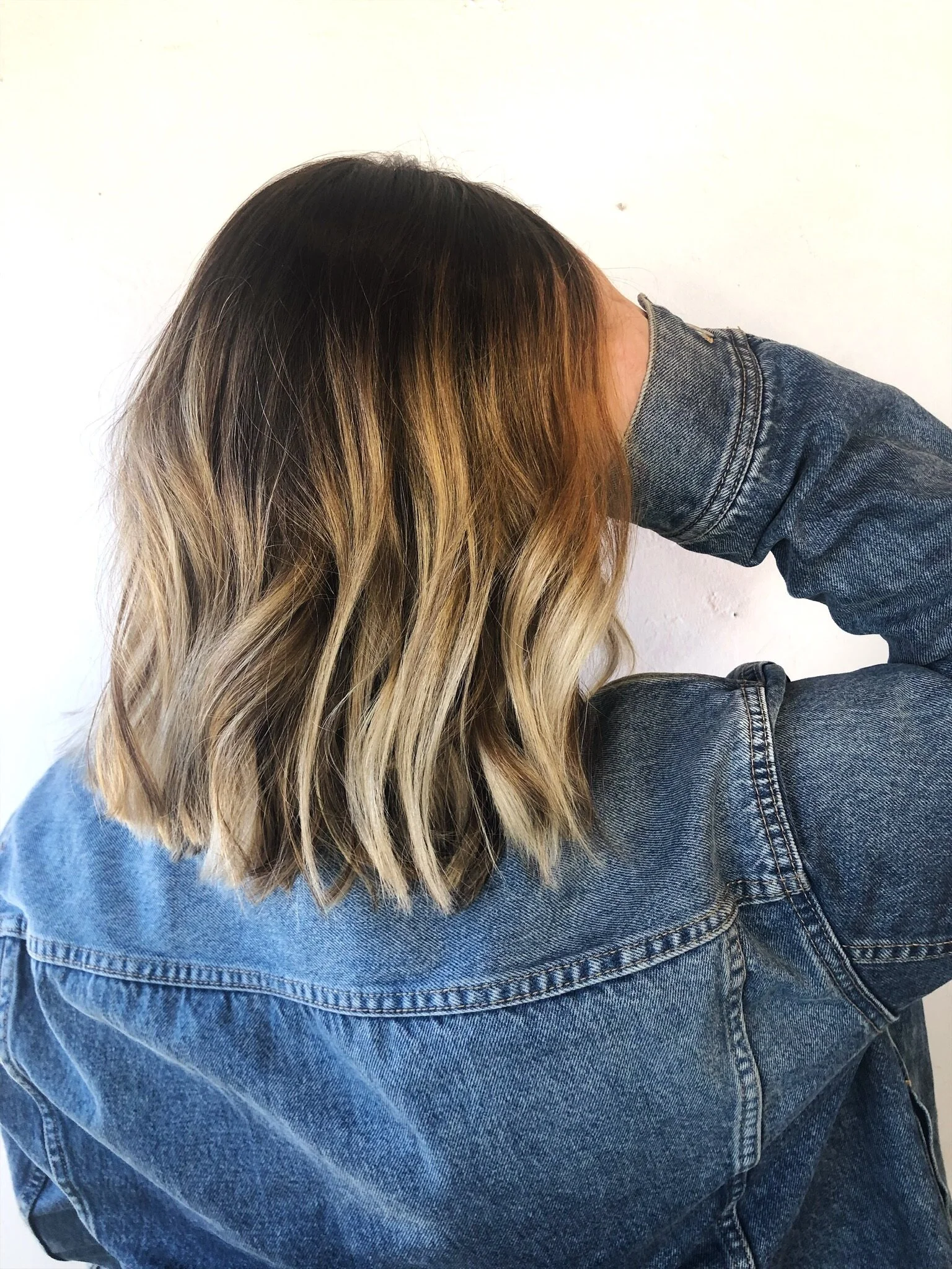 Person with shoulder-length wavy hair, balayage highlights, wearing a denim jacket, leaning forward with head down against a plain white wall.