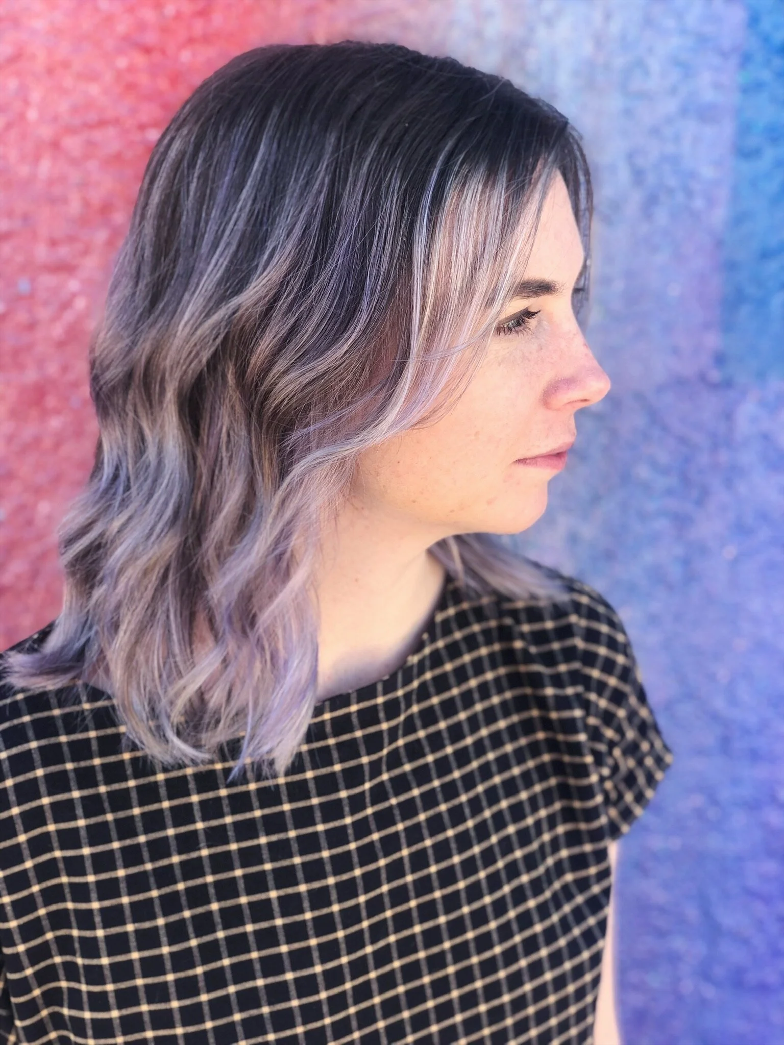 Side profile of a woman with wavy hair that is dark at the roots and light purple at the ends, standing against a multicolored textured background.