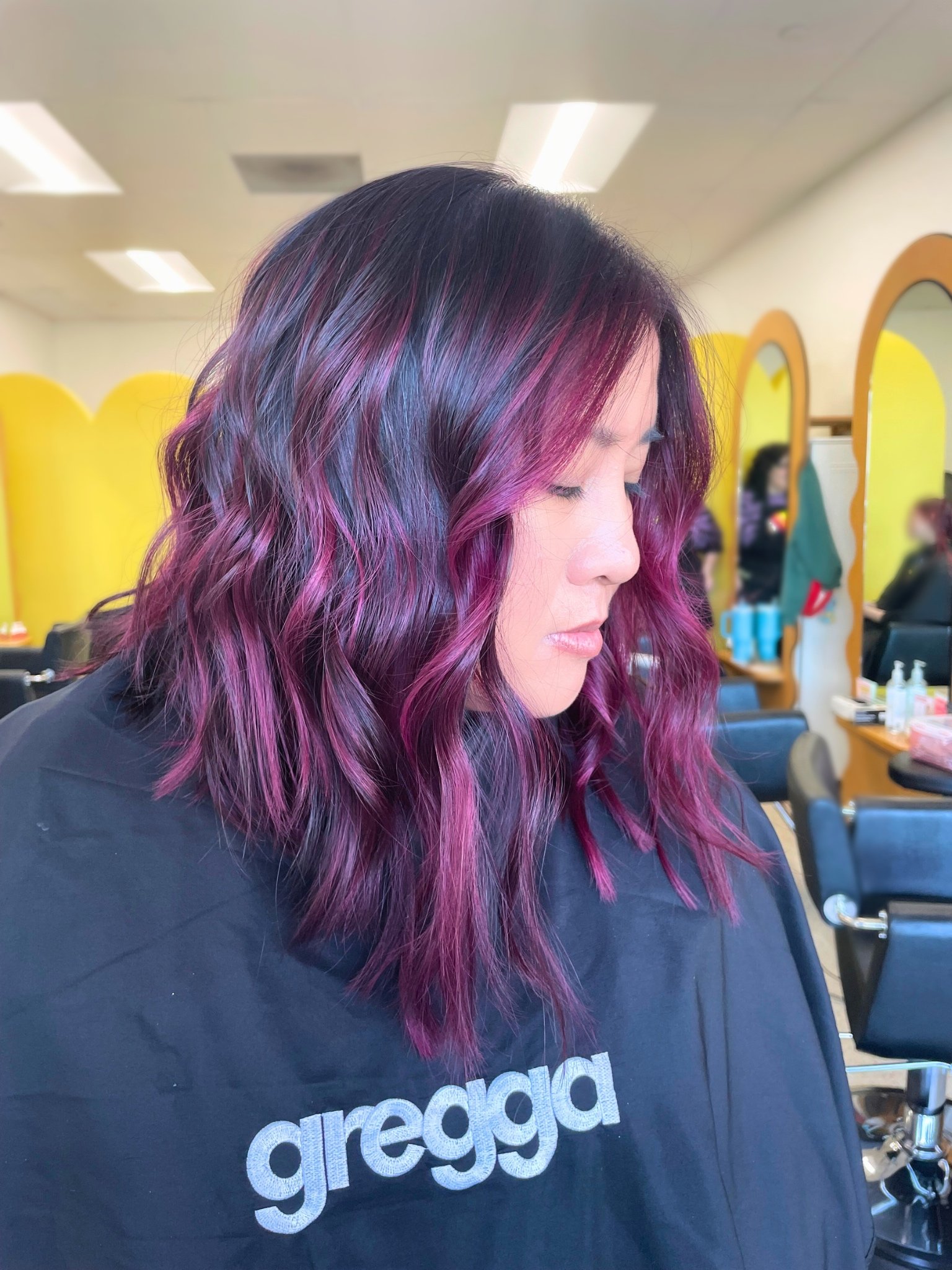 Woman with shoulder-length wavy hair dyed in shades of purple and pink, sitting in a salon.