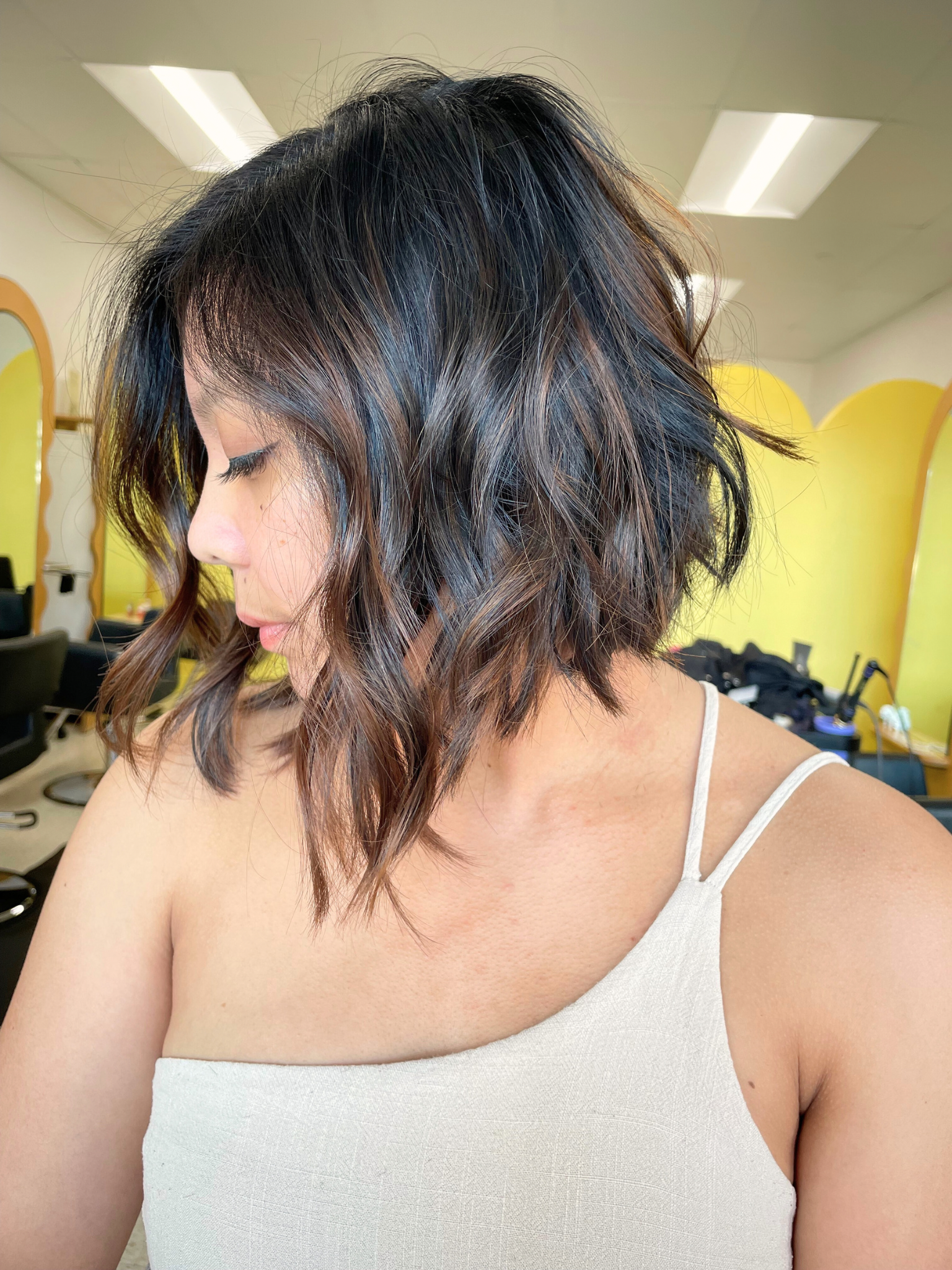 Woman with shoulder-length wavy hair styled in a layered bob, with dark brown and lighter highlights, wearing a white tank top, standing in a salon with yellow dividers in the background.