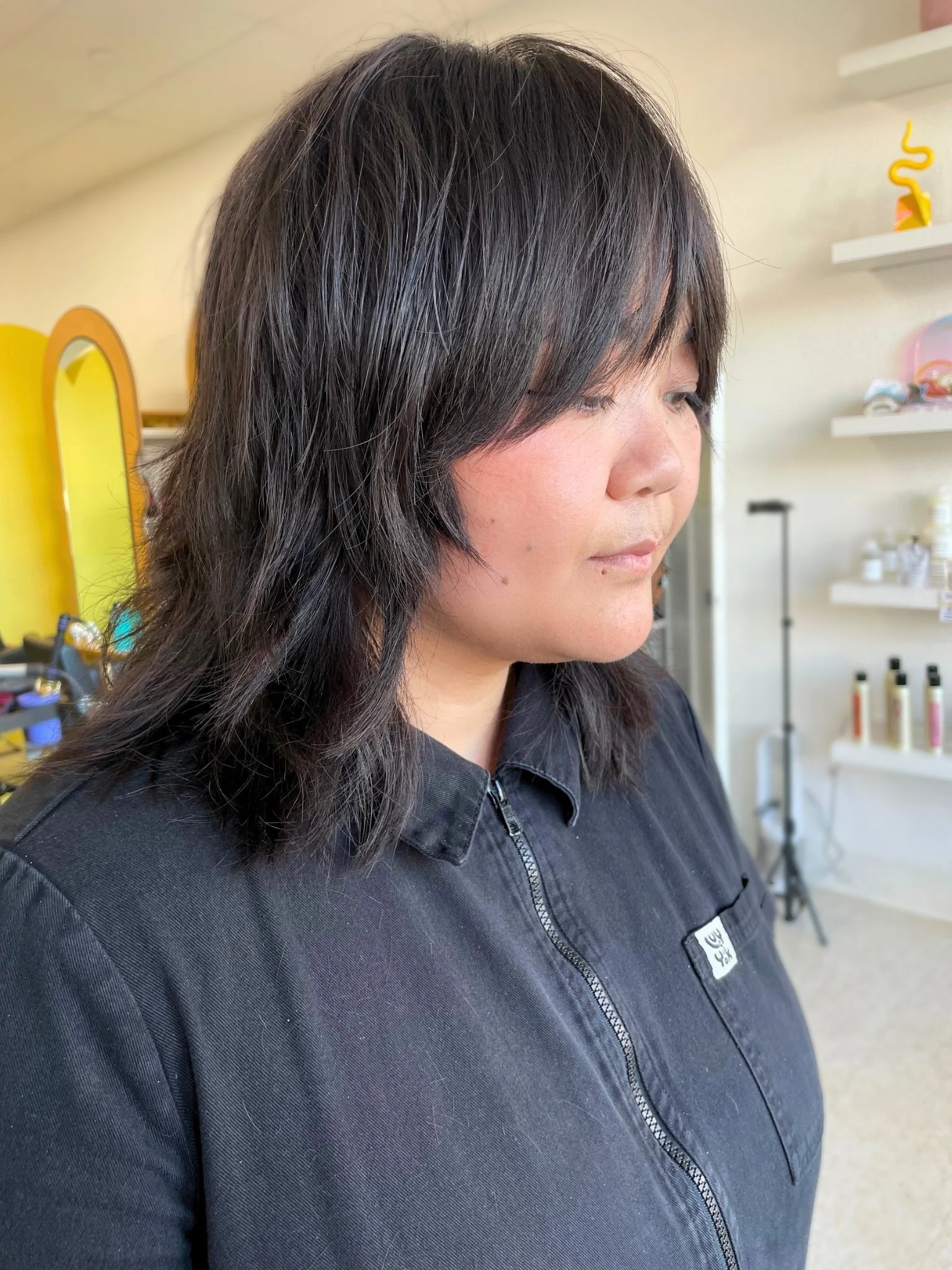 A woman with dark, layered hair wearing a black zip-up jacket standing in a brightly lit room with shelves and products in the background.