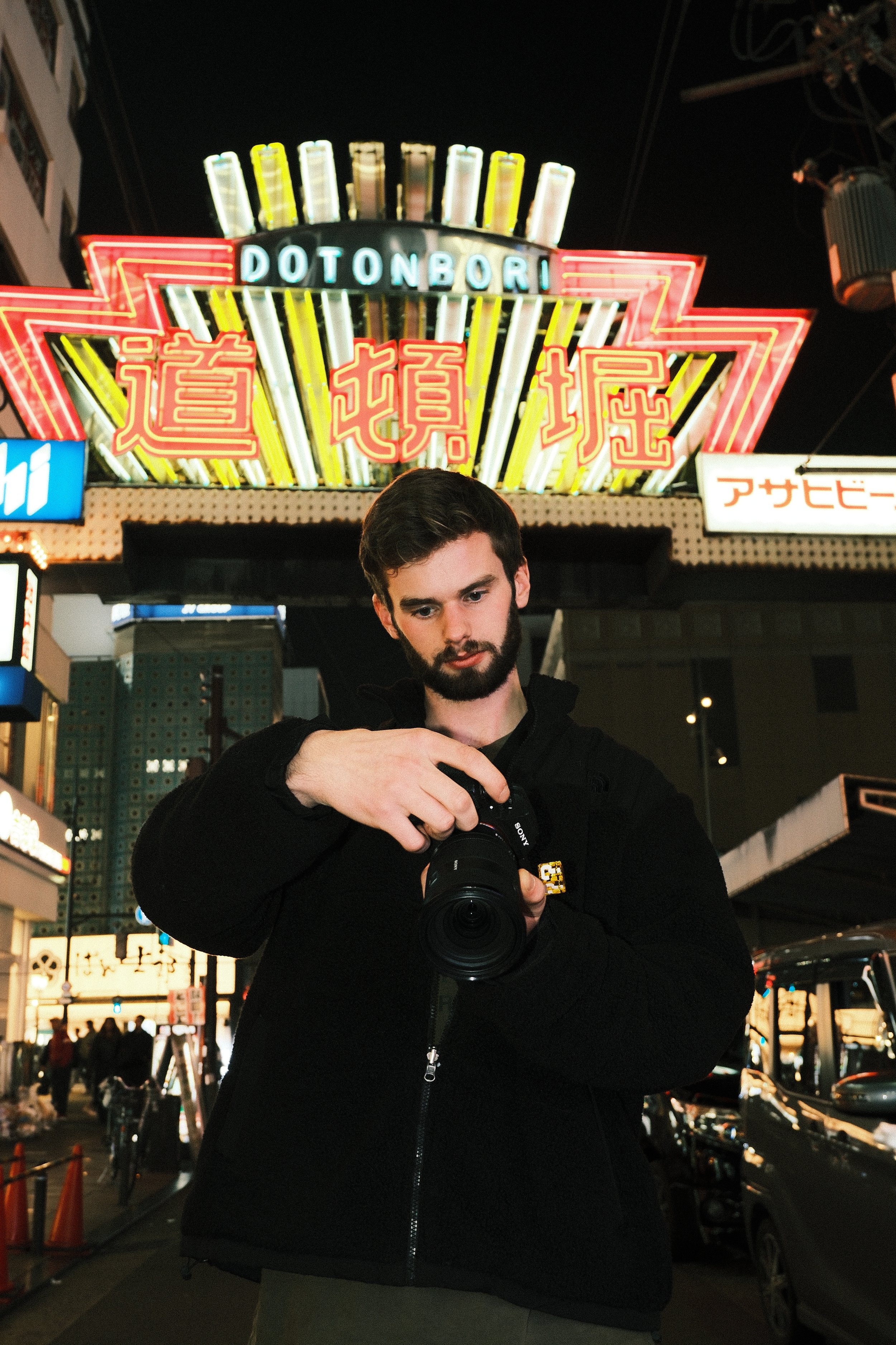 A man with dark hair and a beard holding a camera, standing in a busy city street at night with neon signs and lights.