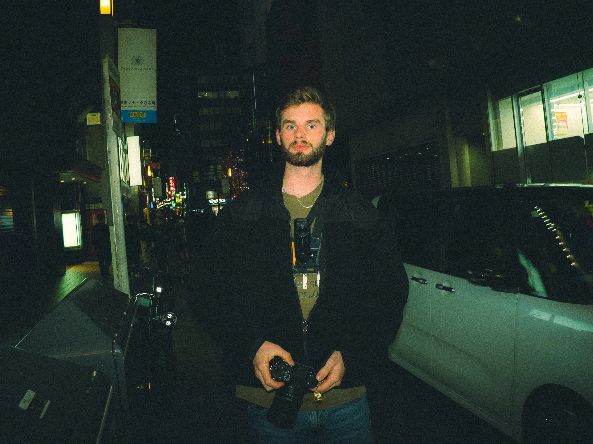 A man with a beard and blue eyes holding a camera, standing next to a white vehicle at night in an urban setting with illuminated signs and buildings in the background.