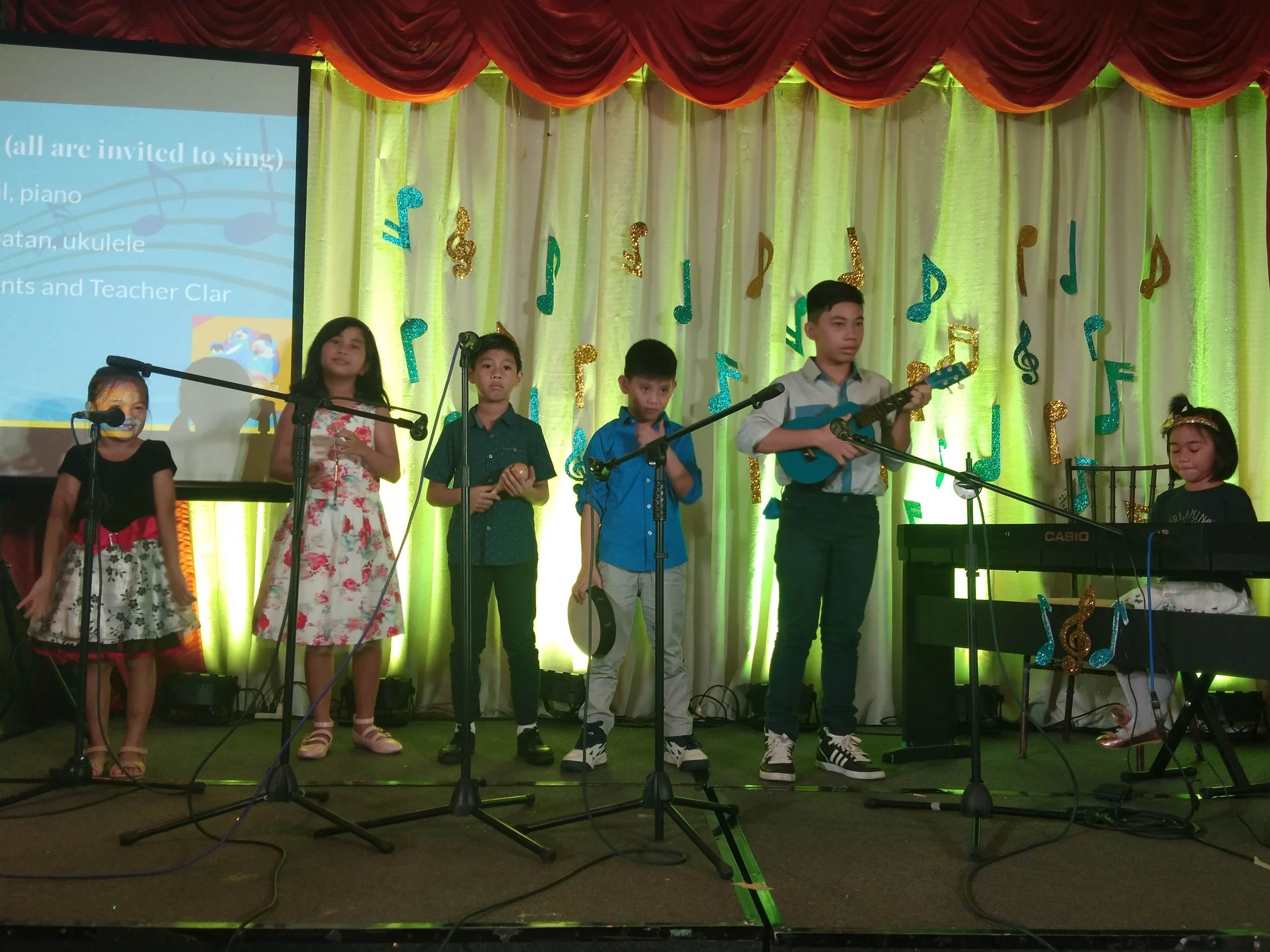 Children performing a musical on stage with green curtains and musical note decorations, one girl playing keyboard, others singing and playing instruments.