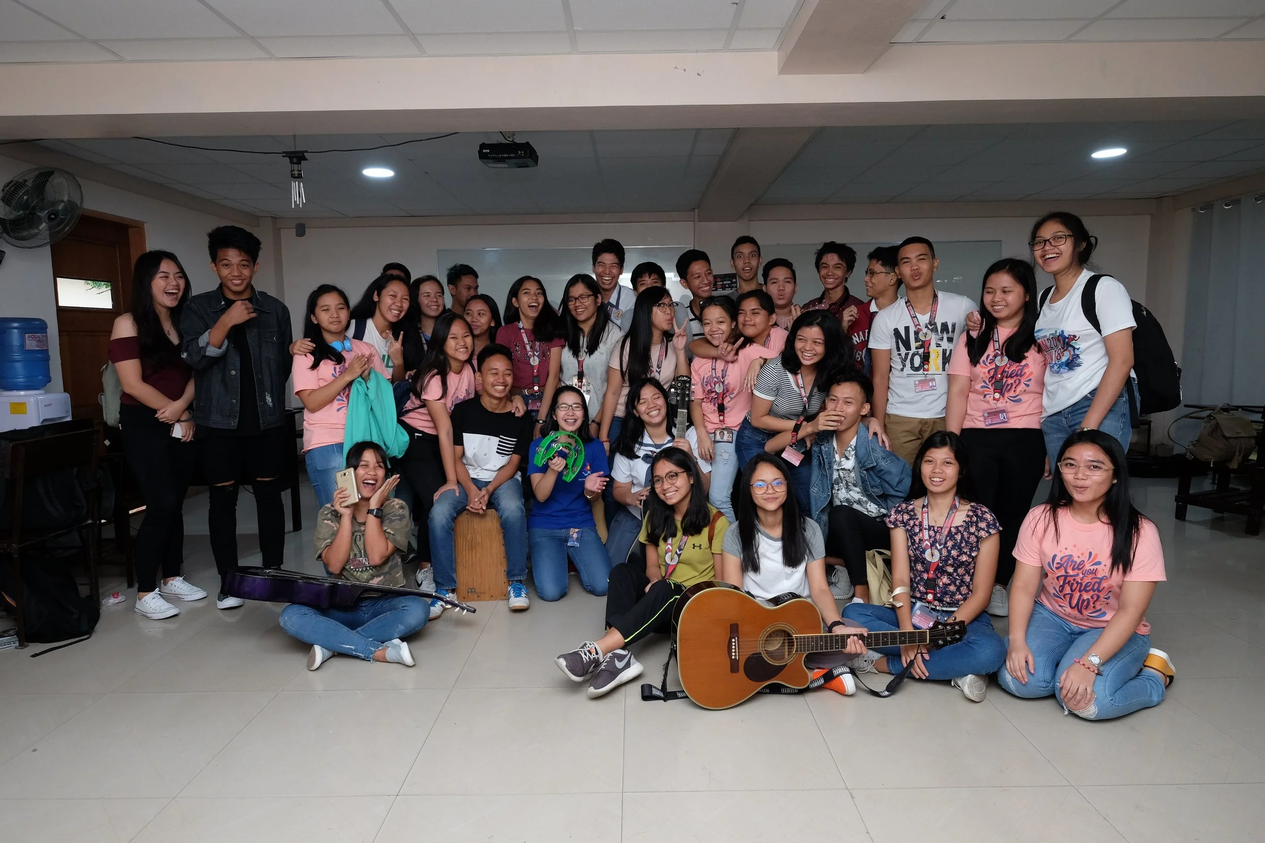 Group of young people smiling and posing for a photo in a classroom or meeting room, some holding musical instruments like guitars and a tambourine.