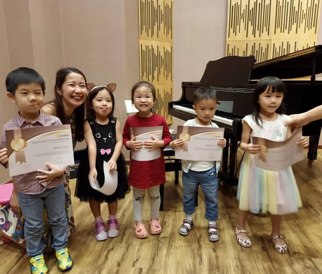 Six children and a woman posing with certificates near a black grand piano in a music room.