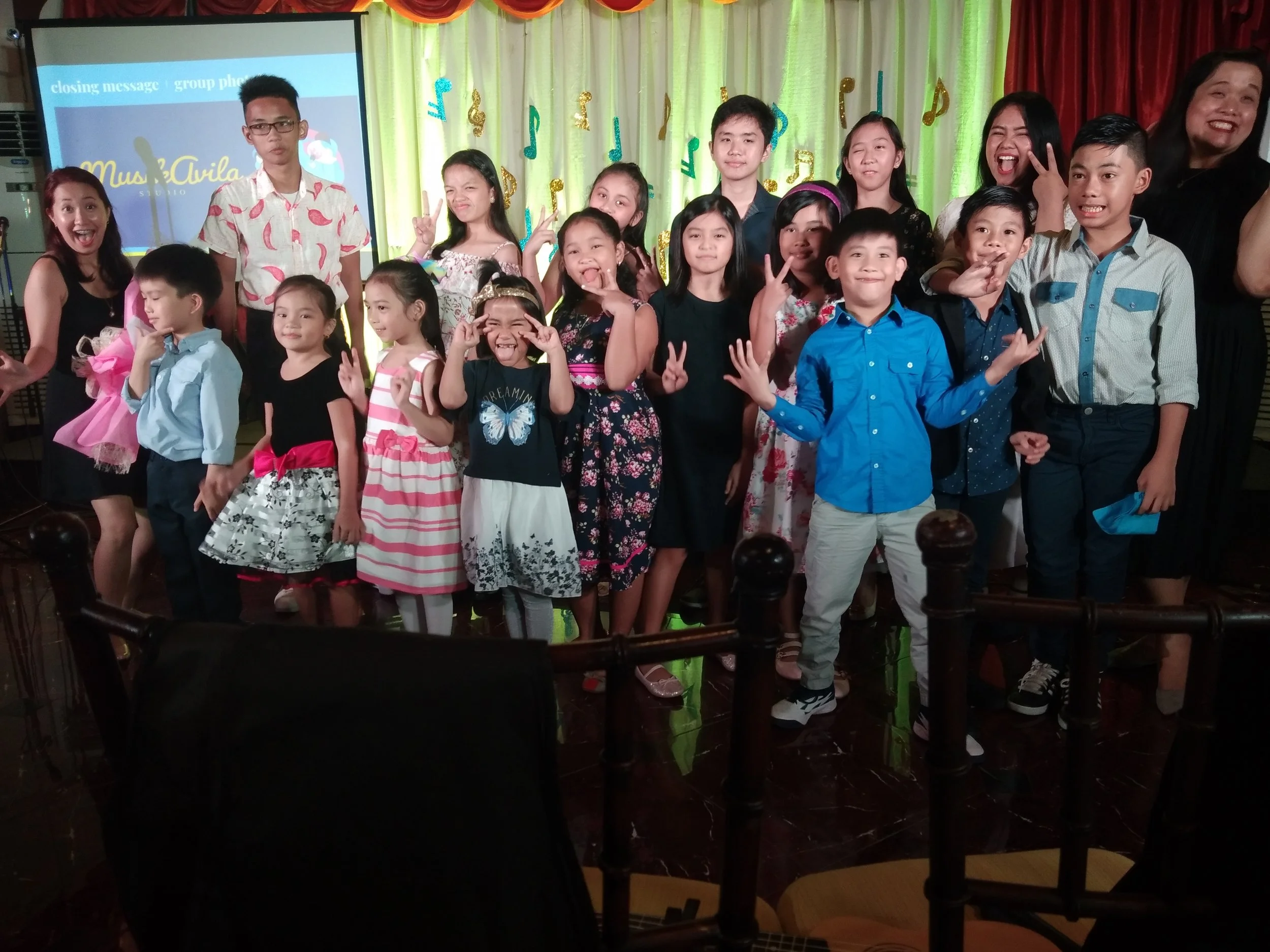 A group of children and adults posing for a photo on a stage decorated with colorful musical notes and drapes, some children making peace signs and smiling.