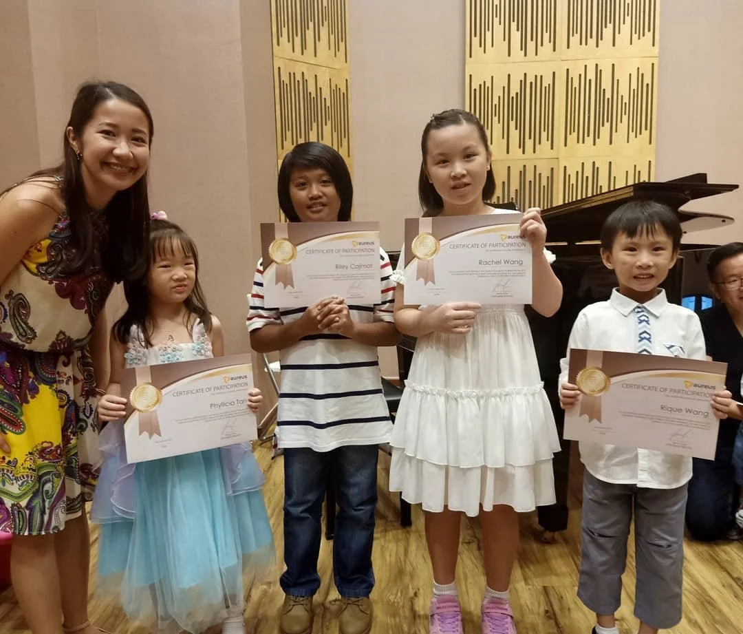 Children holding certificates at a music recital, with a woman smiling on the left.