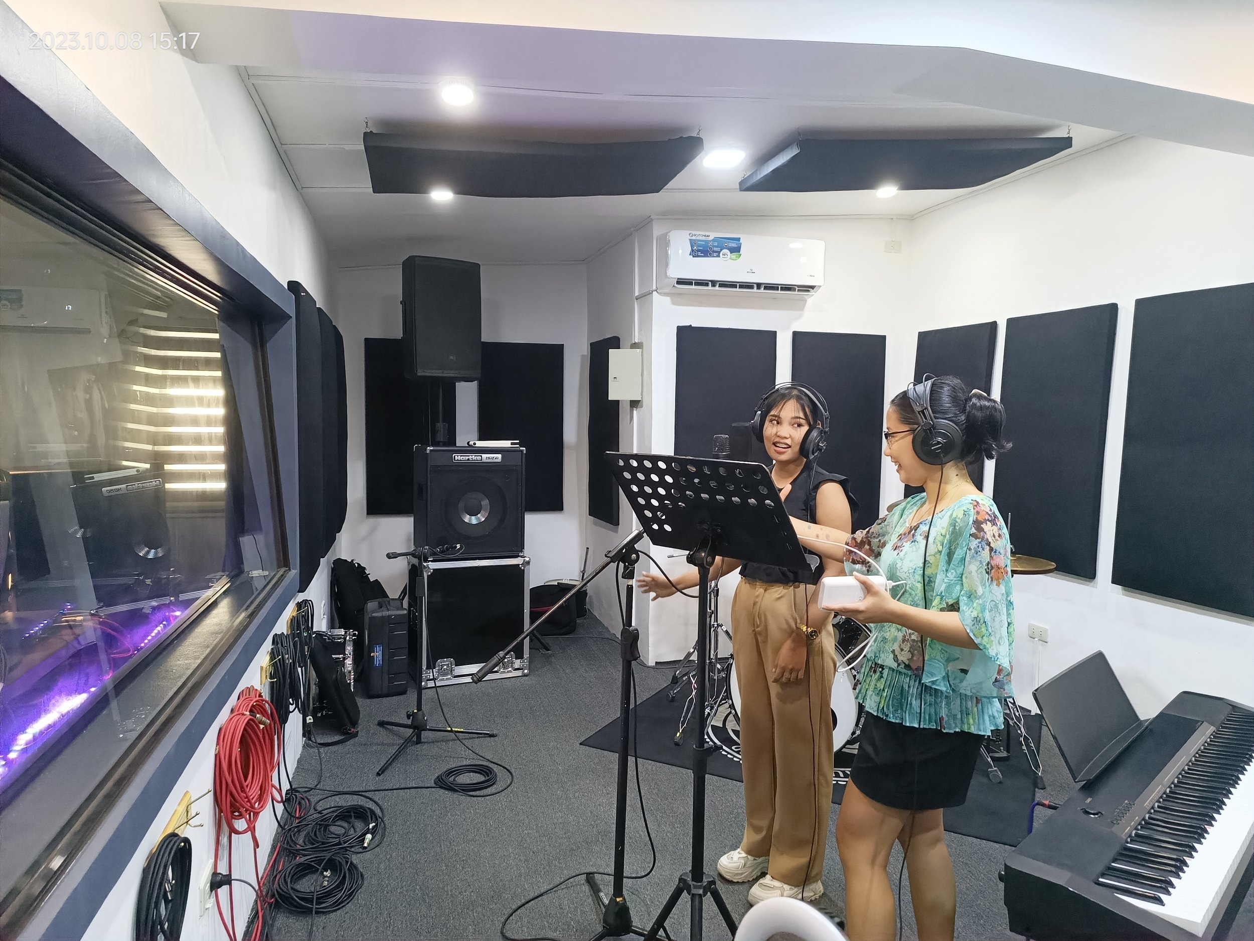 Two women wearing headphones stand in a recording studio with musical equipment, microphones, a keyboard, and soundproofing panels on the walls.