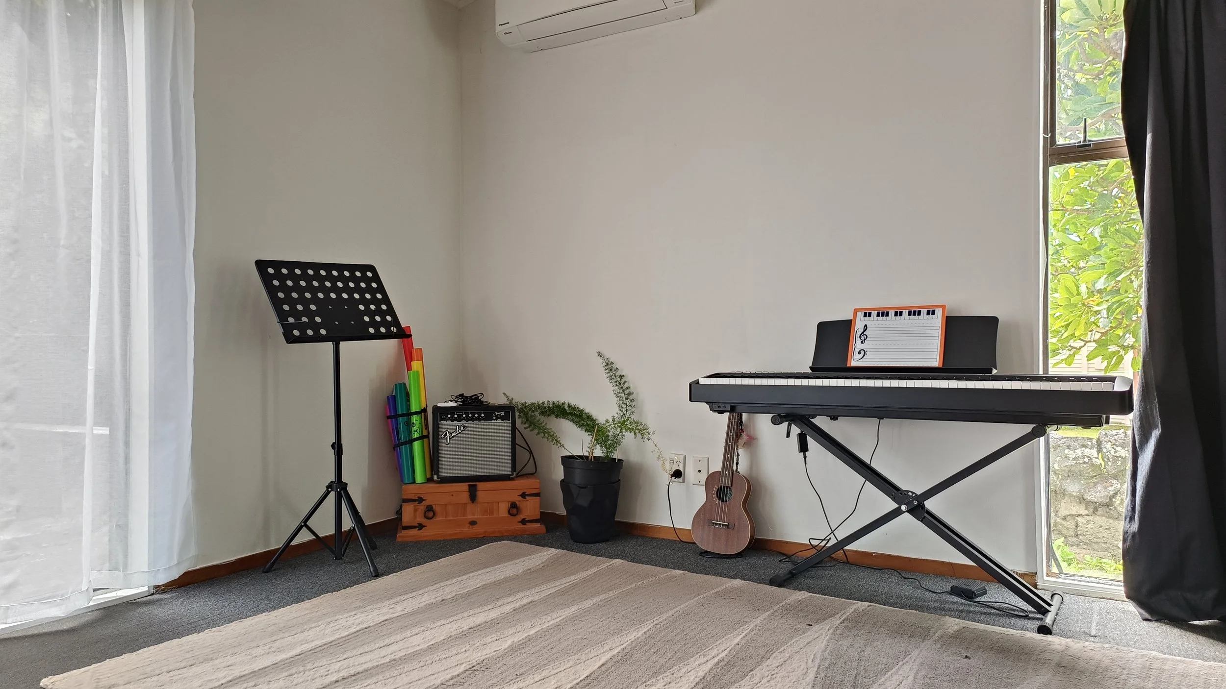 Music room with a digital piano, sheet music, and a guitar, next to a window with green foliage outside.