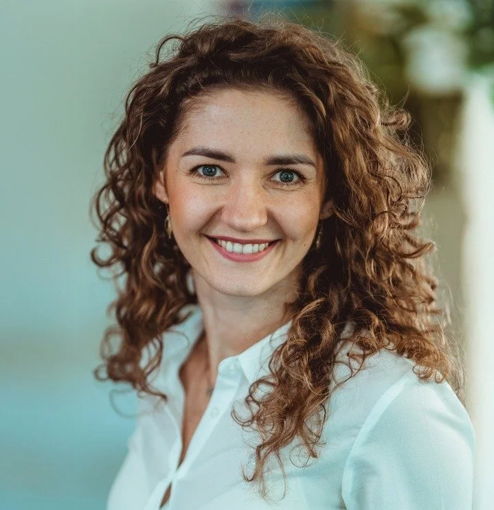 Portret uśmiechniętej kobiety z kręconymi rudymi włosami w białej koszuli na plenerowym tle. Anna Kneba, CEO ESGALL.COM i ekspertka ds. strategii zrównoważonego rozwoju.