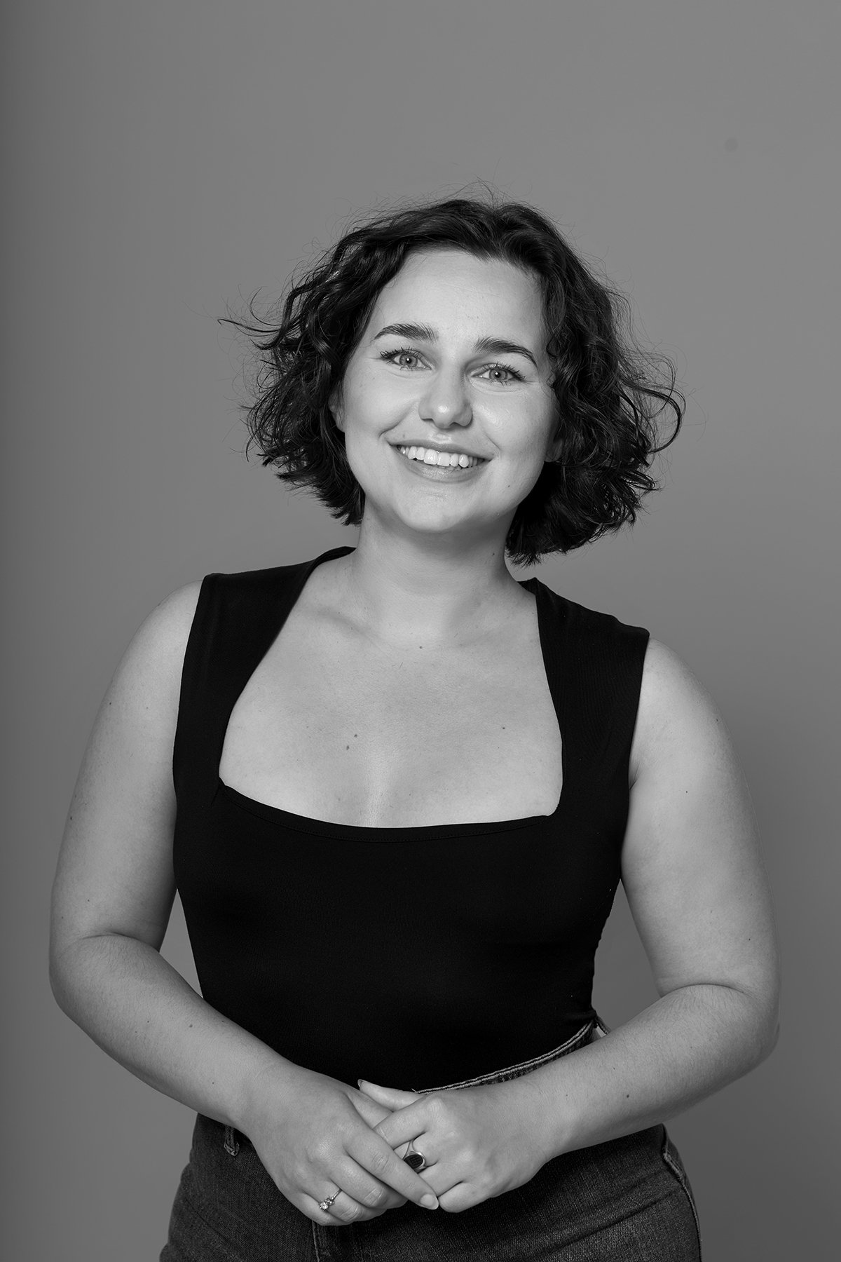 A woman, the author Elisabeth G. Mark, with curly dark hair smiling, wearing a sleeveless black top and dark pants against a plain background.