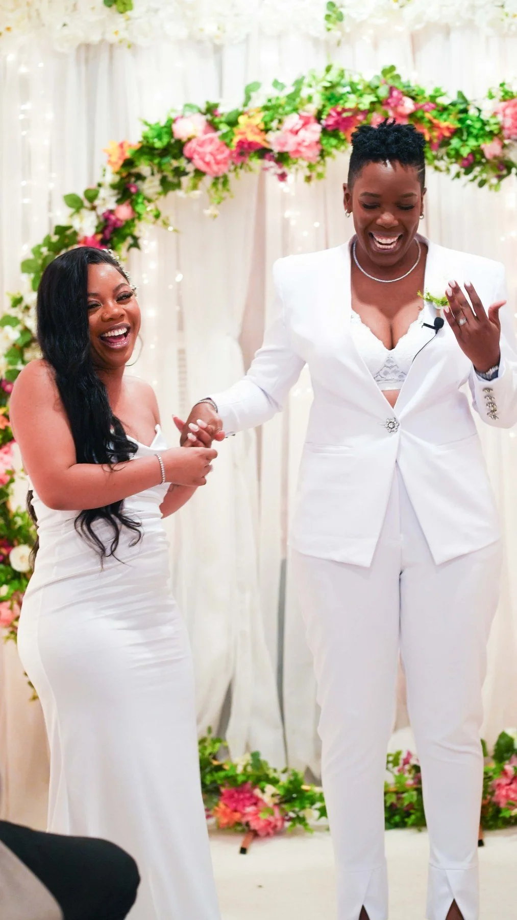 Couple in white laughing and holding hands in front of a floral arch during their New York City wedding ceremony, one partner showing off her ring