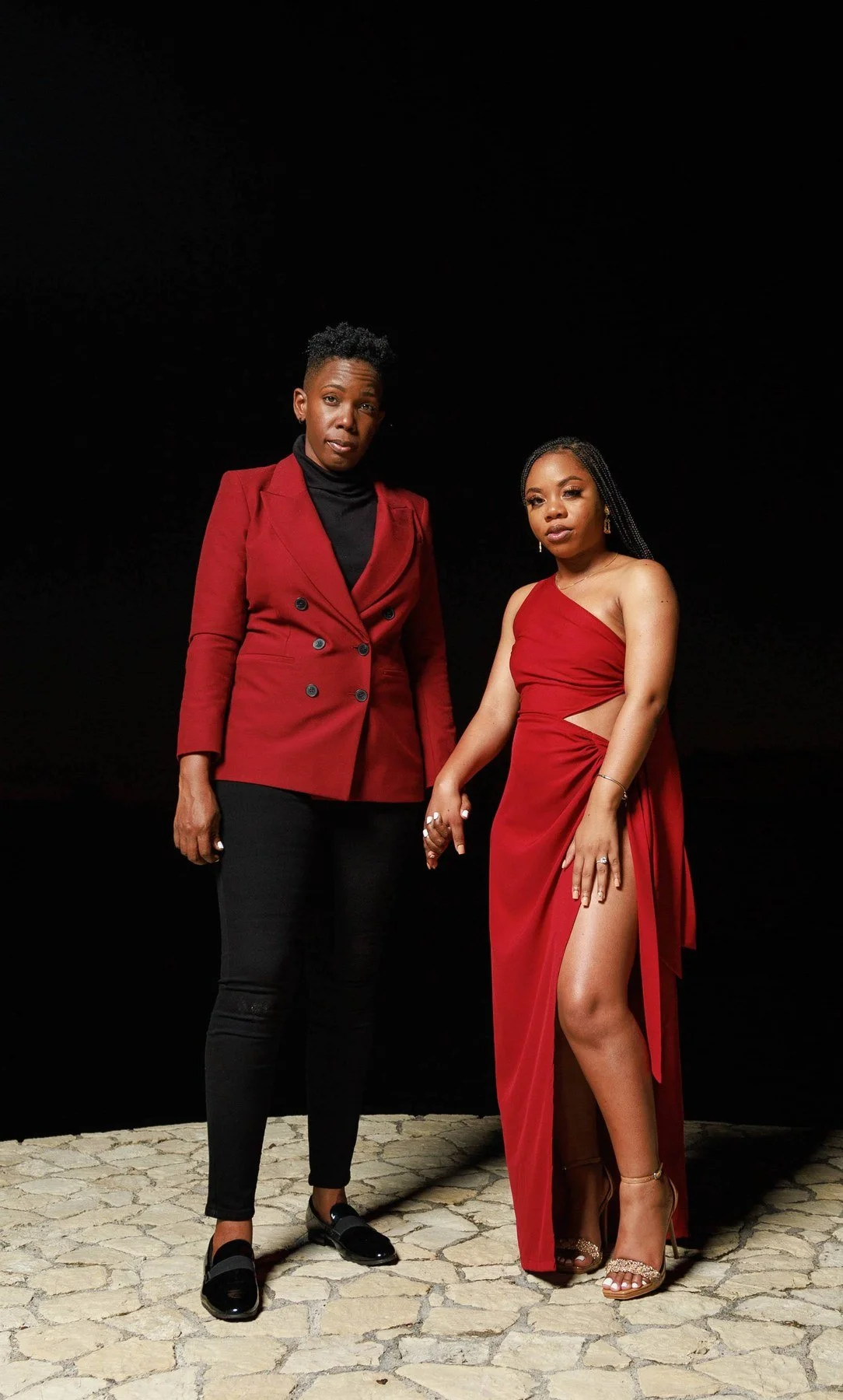 Editorial engagement portrait in Jamaica of a couple holding hands on a stone patio at night, one in a red double-breasted blazer and black turtleneck, the other in a one-shoulder red gown with a high slit