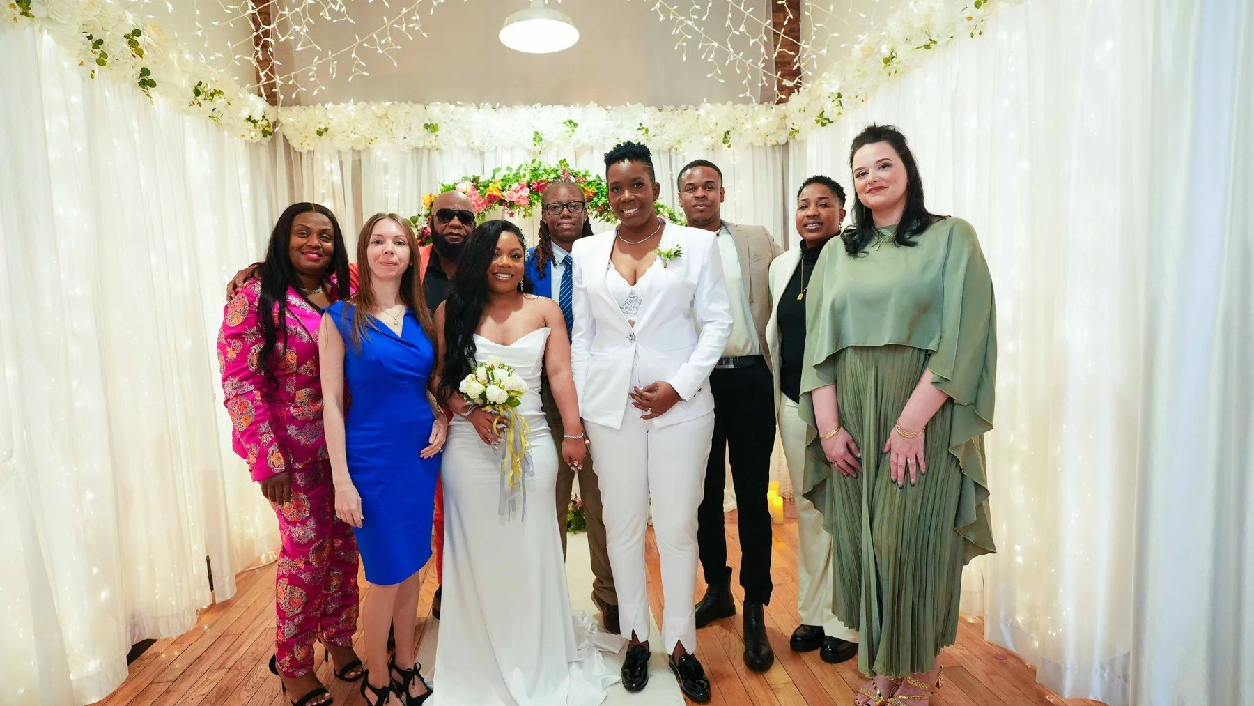 Newlyweds in white standing with family and friends under a draped canopy with string lights and a floral arch after their New York City wedding ceremony