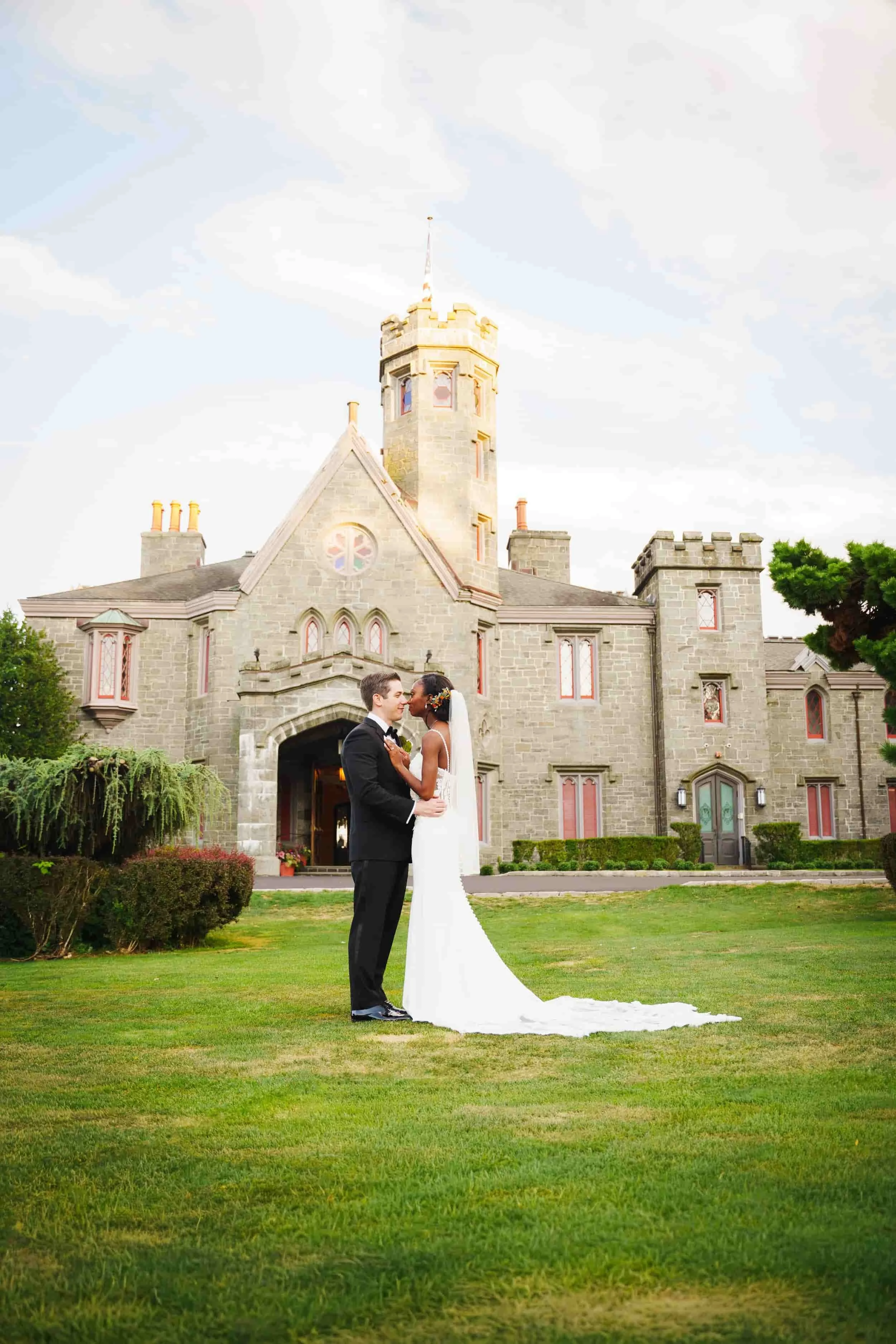 A bride and groom standing close together on a lush green lawn in front of Whitby Castle with multiple turrets and red-trimmed windows, during their wedding photoshoot.