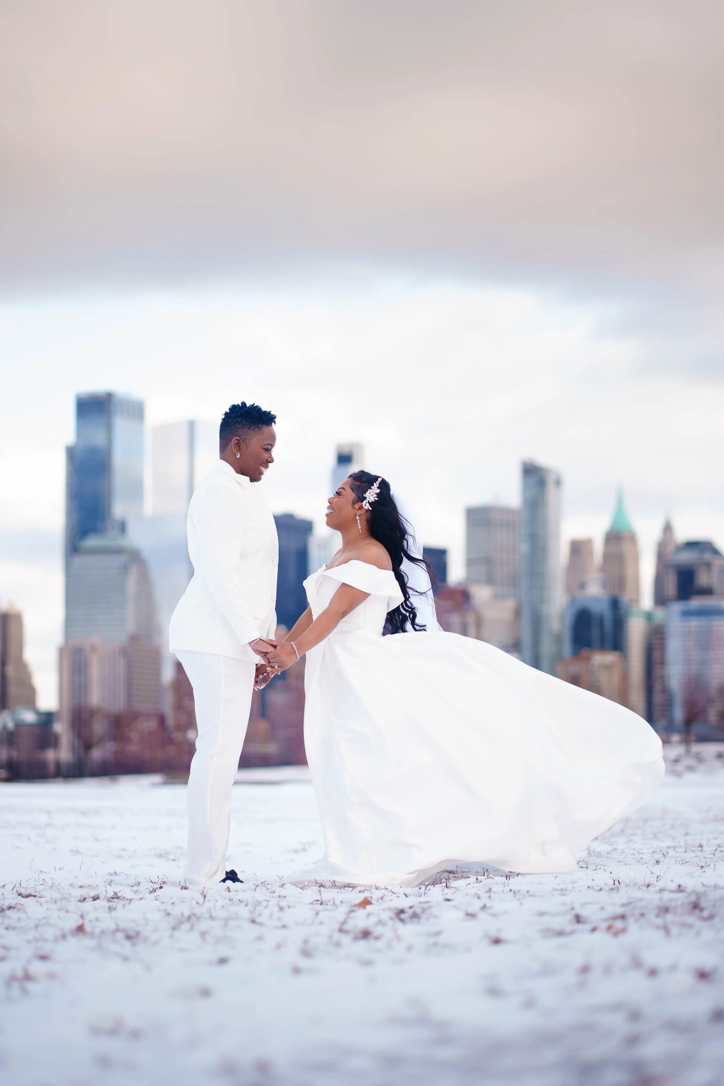 A couple dressed in white wedding attire holding hands on a snowy field with a city skyline in the background at Liberty State Park
