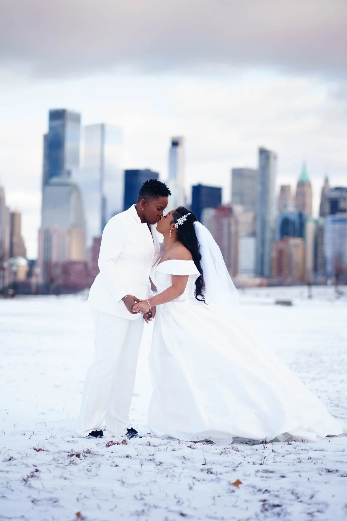 Winter LGBTQ wedding portrait at Liberty State Park in Jersey City, the couple kissing and holding hands in the snow as a veil and gown sweep behind them