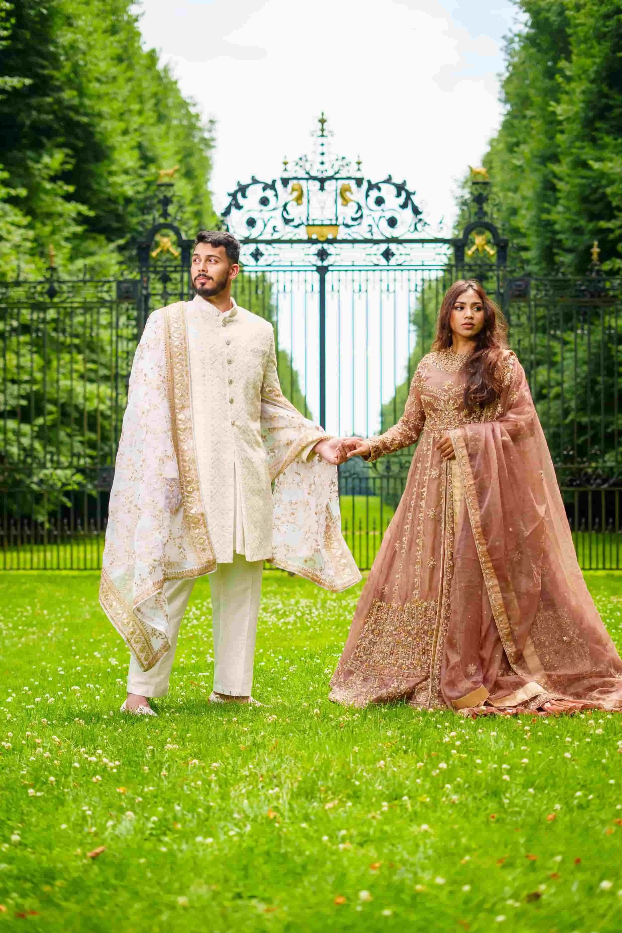 A couple wearing traditional Indian wedding attire holding hands in front of a decorative iron gate and lush green trees at Old Westbury Gardens.