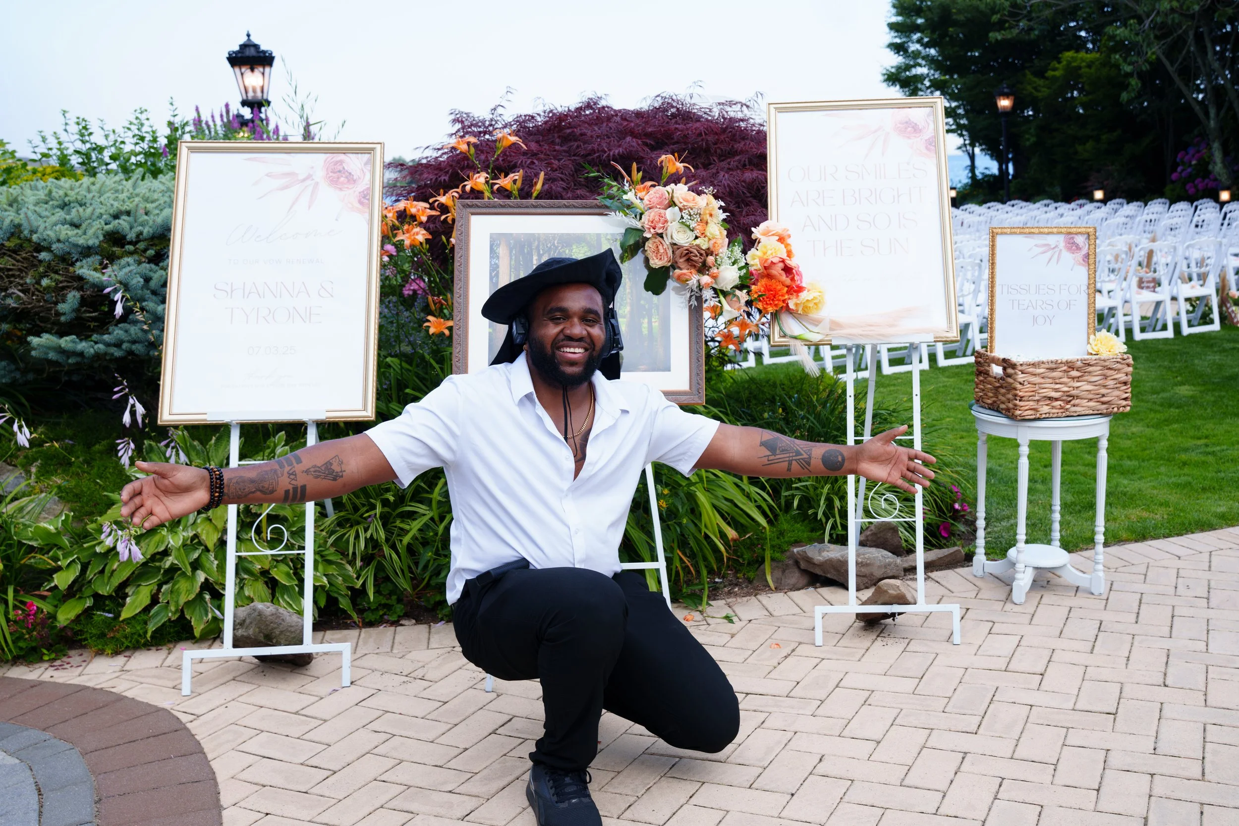 New York City Wedding Videographer Dane Nelson smiling and gesturing with open arms in front of decorative signs and flower arrangements at an outdoor event at Riveria Mansion in Long Island, with white chairs and lanterns in the background.