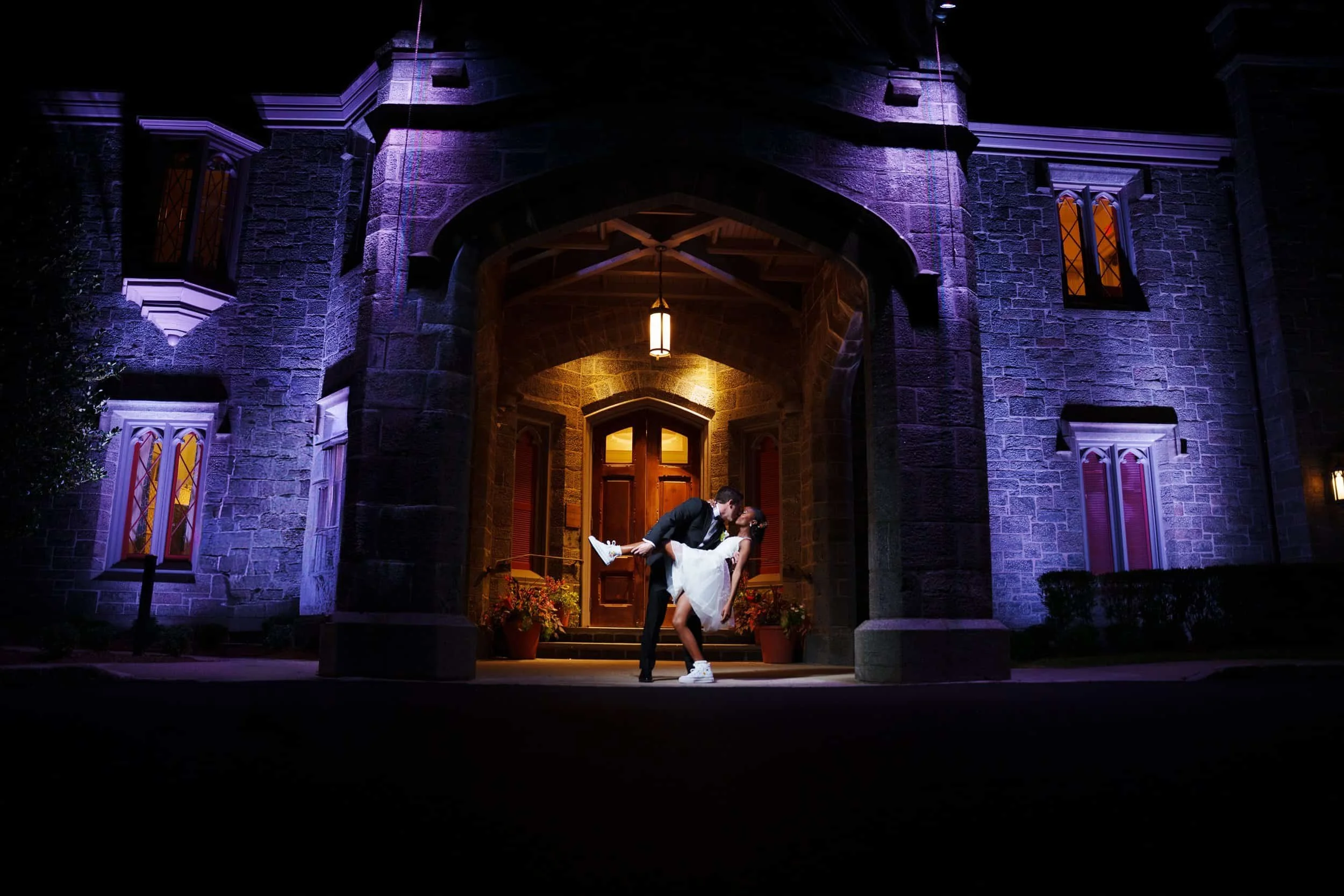 A couple dressed in wedding attire sharing a kiss beneath a stone archway at night at Whitby Castle in Long Island, illuminated by warm yellow and purple lighting outside a large stone building with windows and potted plants.