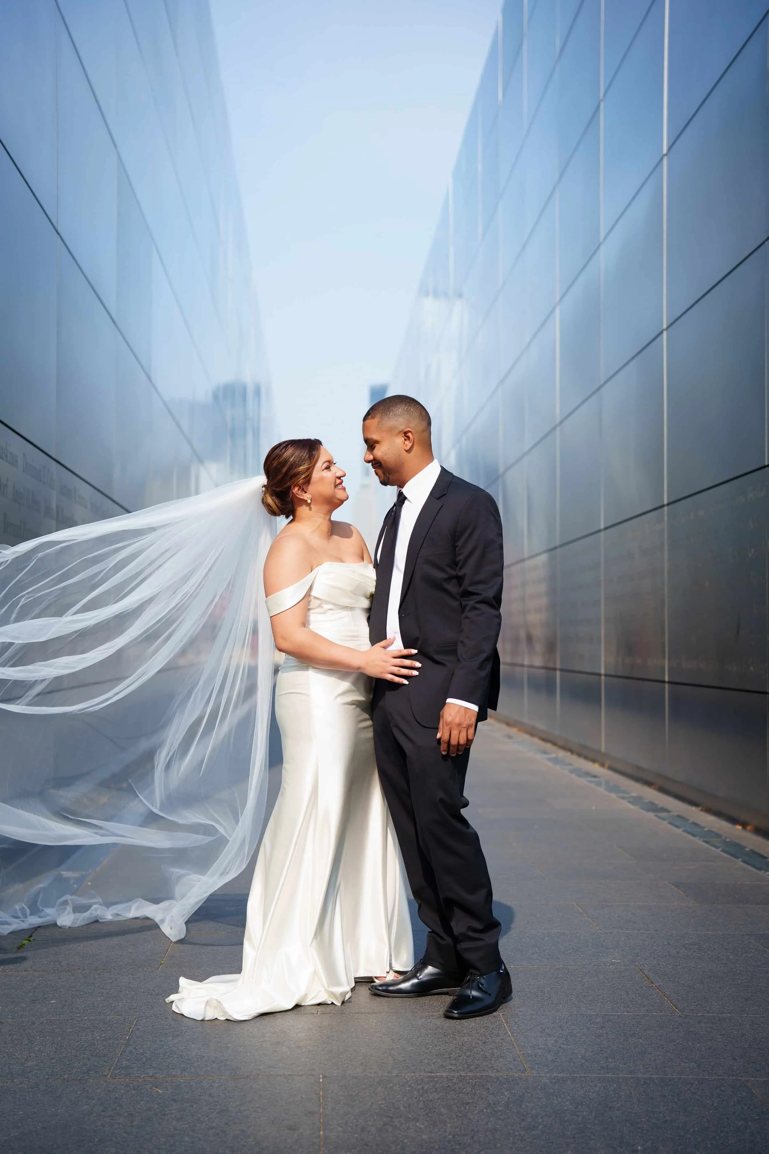 A bride and groom in wedding attire standing close together outdoors, smiling and looking into each other's eyes,  at 9/11 memorial at liberty state park