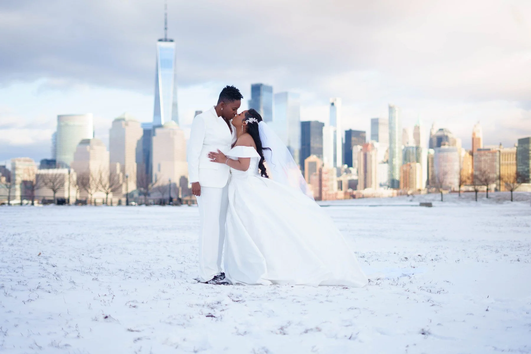 Couple kissing in a snowy field at Liberty State Park, one in a white suit and one in a white off-the-shoulder gown and veil, with One World Trade Center and the Manhattan skyline behind them