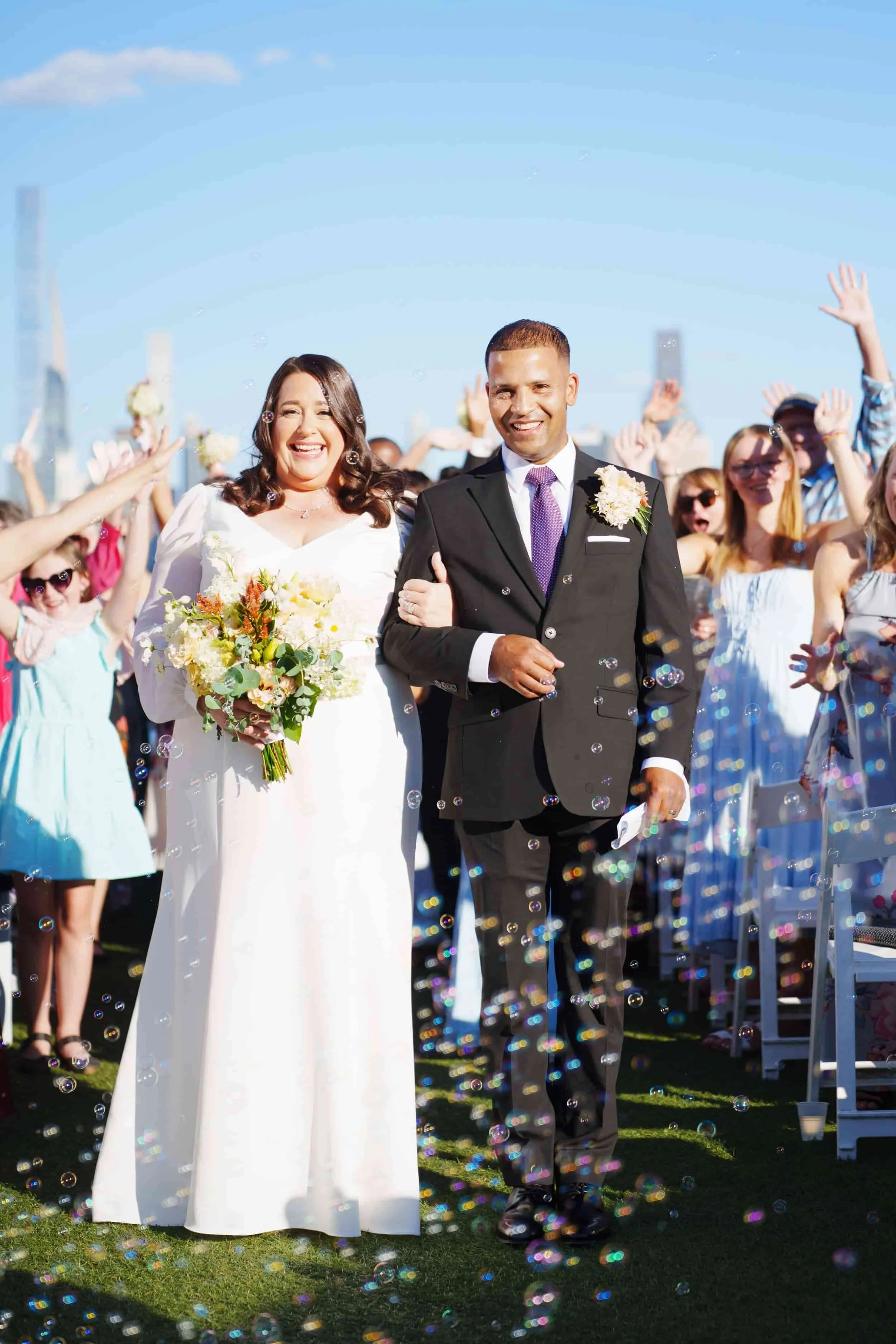 A newlywed couple walks arm-in-arm through a crowd of wedding guests outdoors at the Envue Hotel, in New Jersey, on a sunny day, with bubbles in the air and a city skyline in the background.
