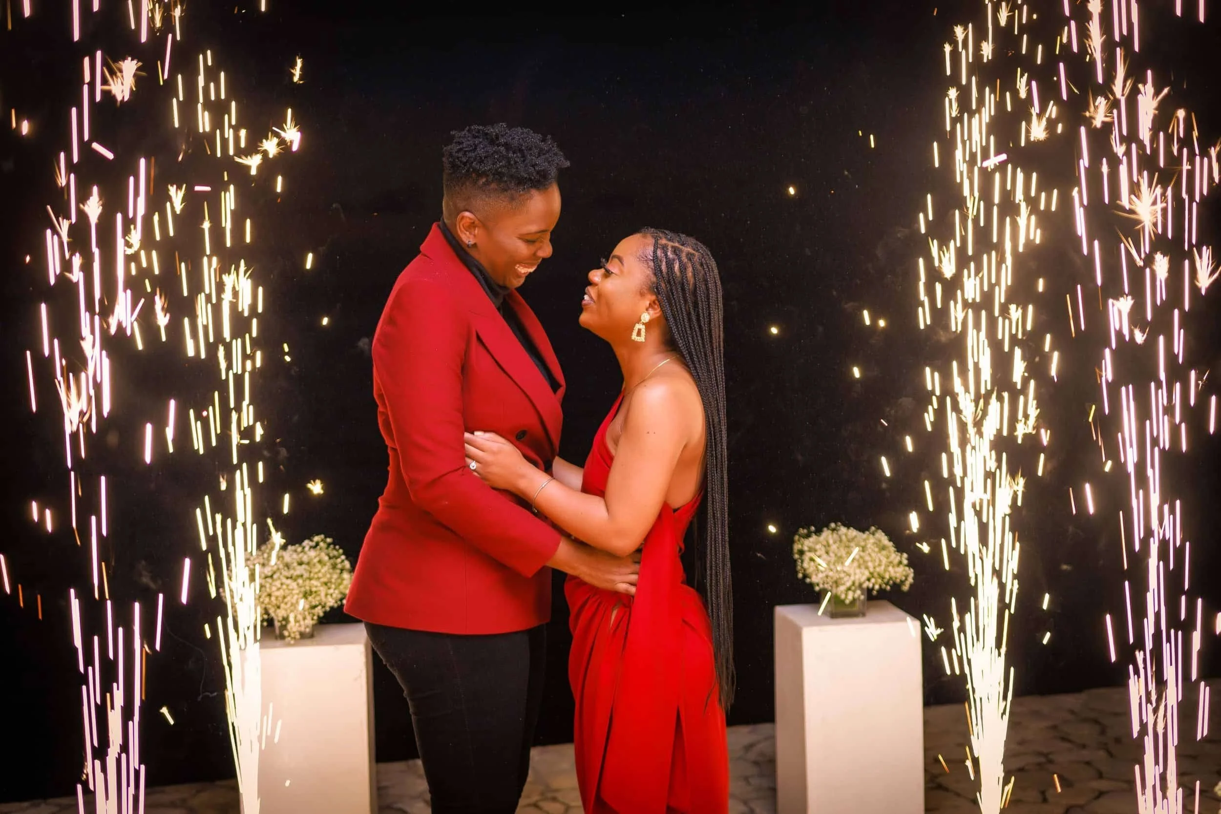 Two women in elegant red outfits embrace with sparklers in the background.